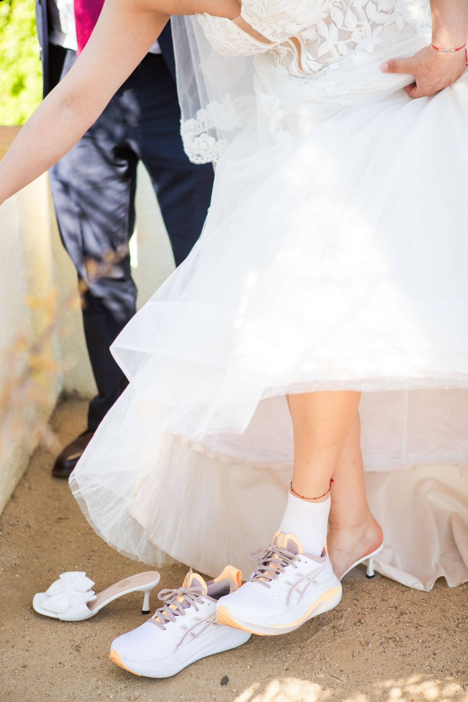 Bride in a white lace wedding dress changes from white high heels into comfortable white sneakers outdoors on a dirt path.