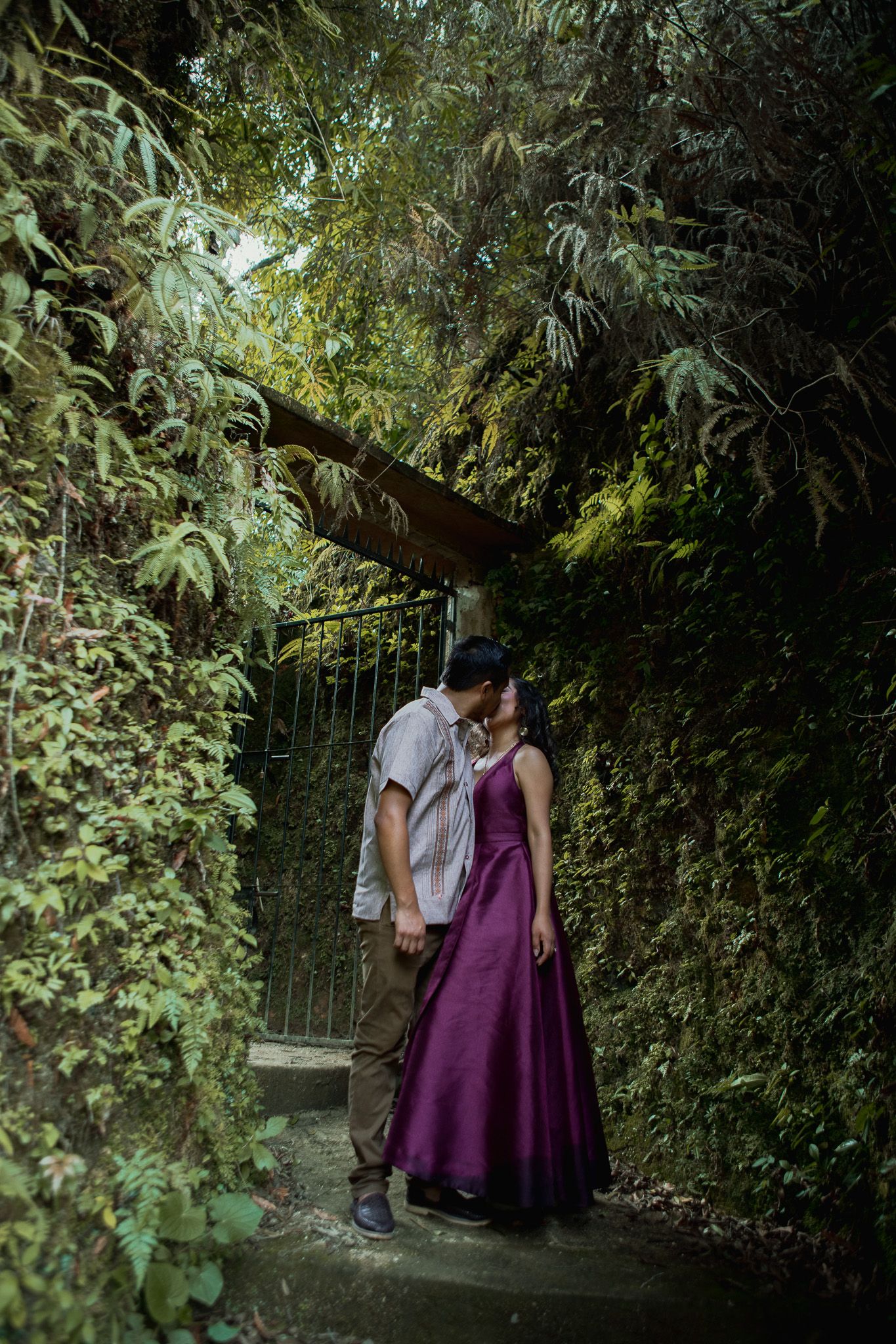 Couple in a purple gown and gray shirt kiss beneath a wooden gate archway surrounded by lush green foliage on a stone path.