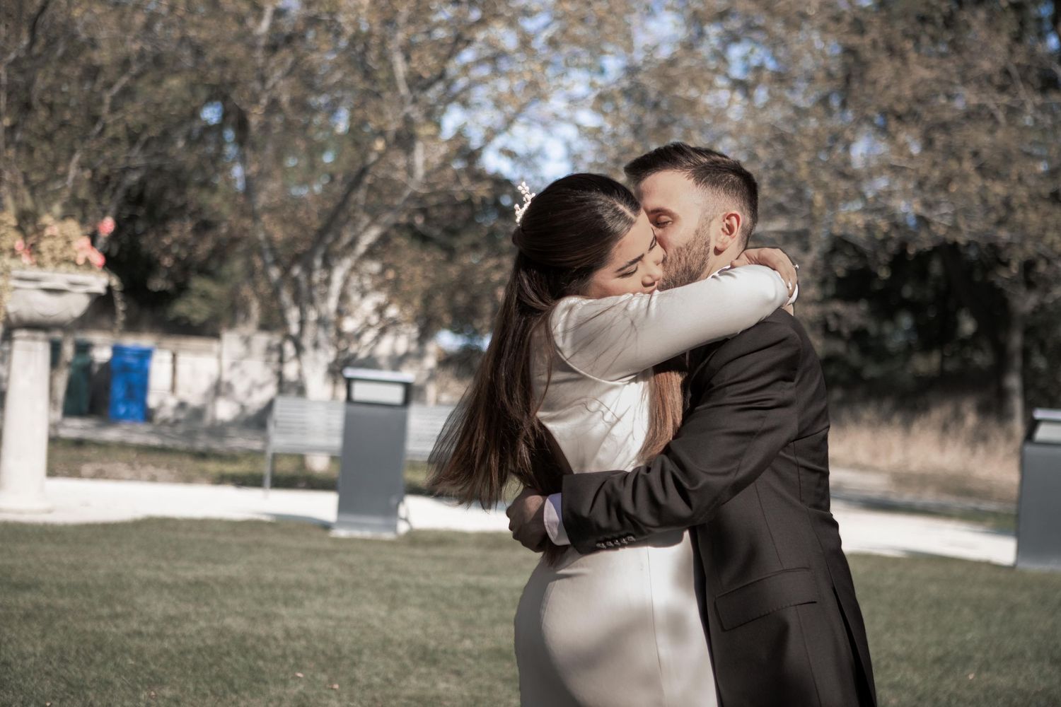 Bride and groom share a romantic kiss outdoors on a grassy lawn surrounded by autumn trees.