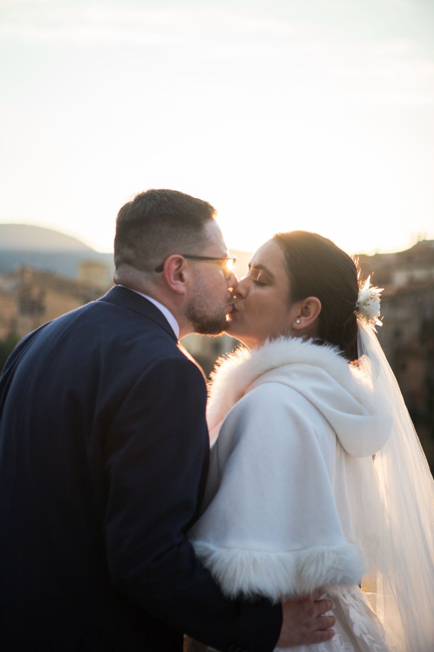 A bride in a white fur-trimmed wrap and veil kisses her groom in a navy suit and glasses during a golden sunset wedding portrait.