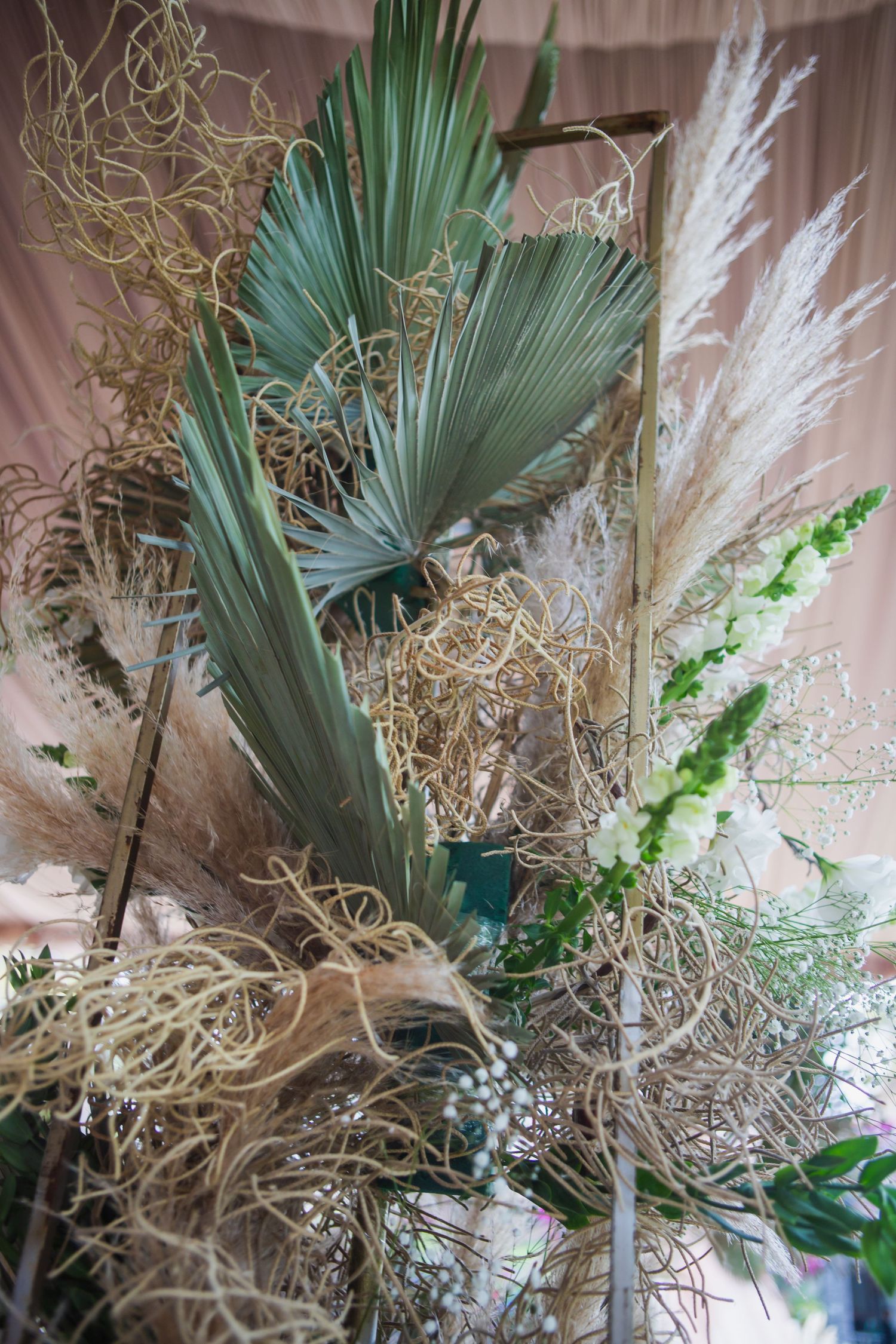 Boho wedding floral arrangement with dried palm fronds, pampas grass, white blooms and twisted natural vines on a wooden frame.