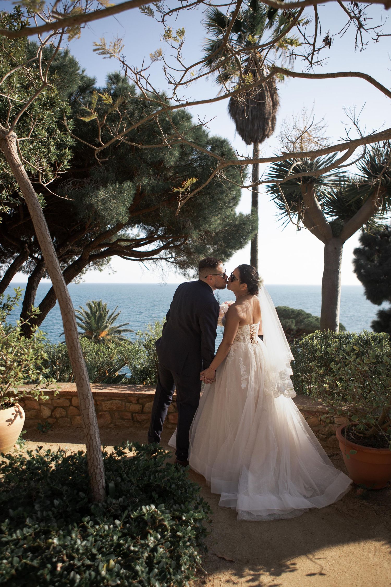 Bride in a flowing tulle gown and veil kisses her groom in a dark suit in a seaside garden with palm trees and ocean views.