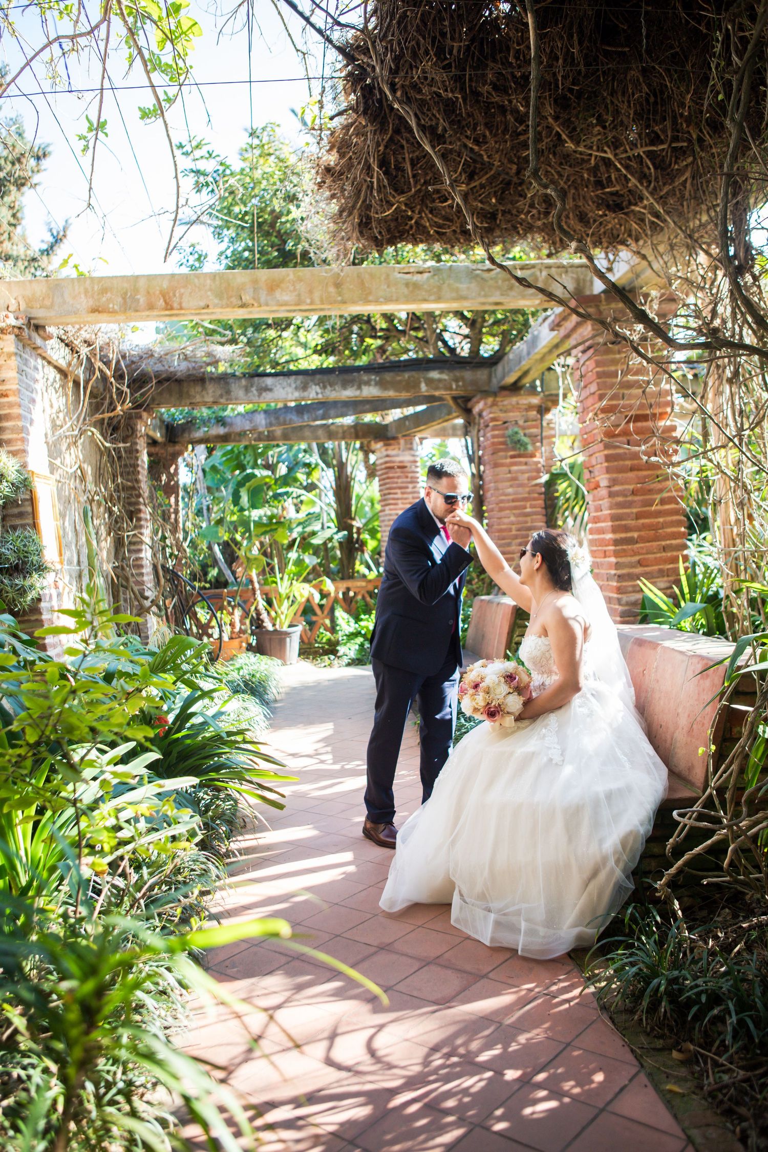 Groom in a dark suit kisses the hand of a bride in a white gown holding a bouquet in a sunlit garden with a brick pergola.