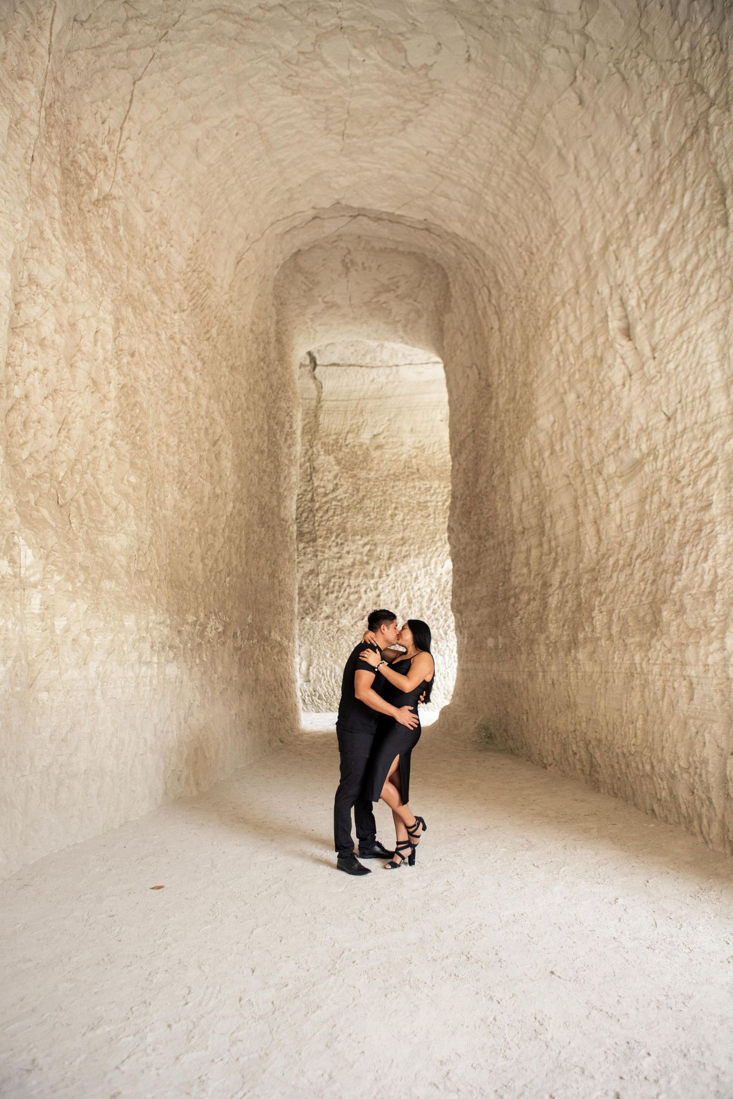 Couple in black outfits share a romantic kiss inside a carved white limestone cave passage, ideal for engagement or wedding photography.