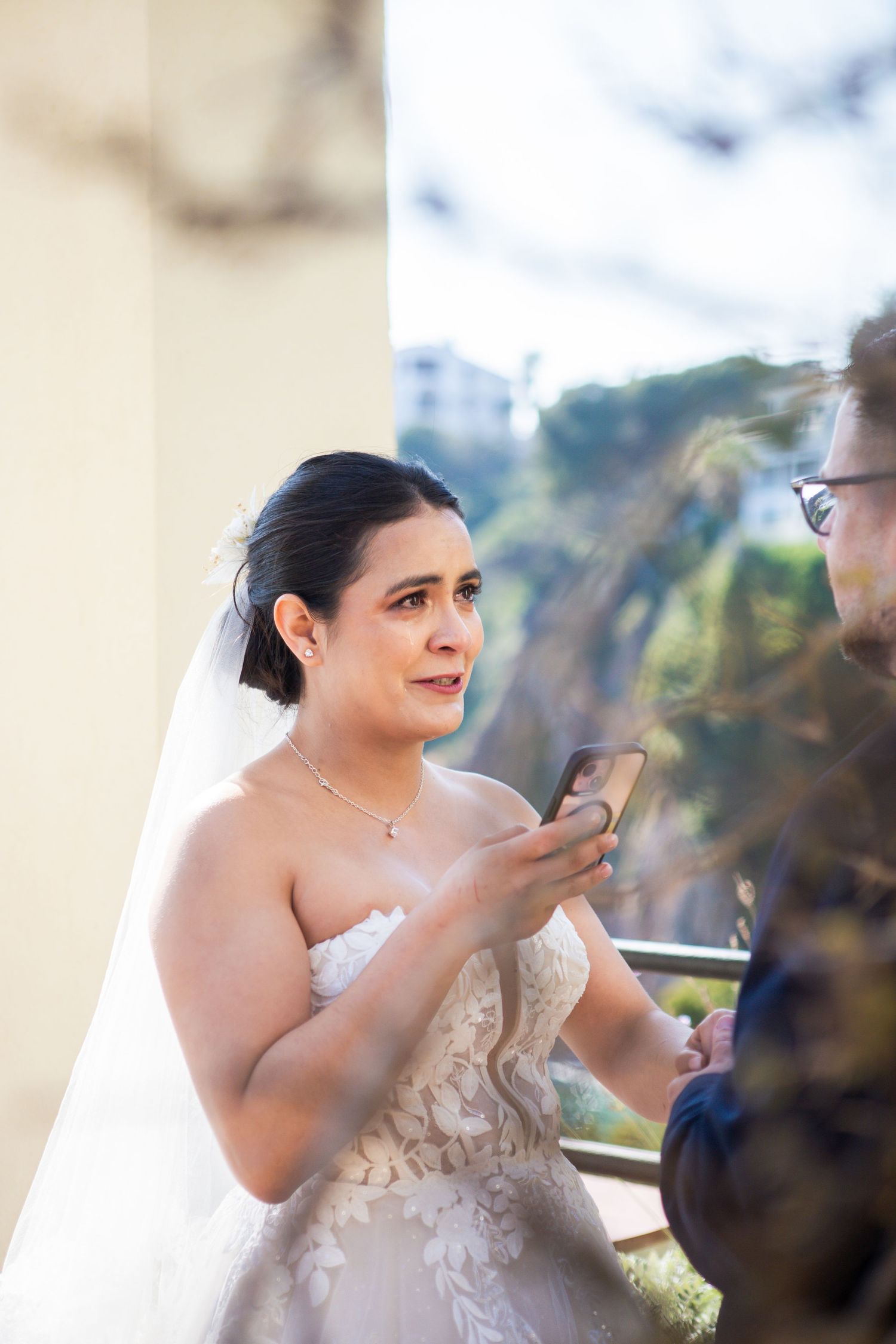 Emotional bride in a strapless lace wedding gown and veil reads vows from her phone during an outdoor ceremony with a hillside view.