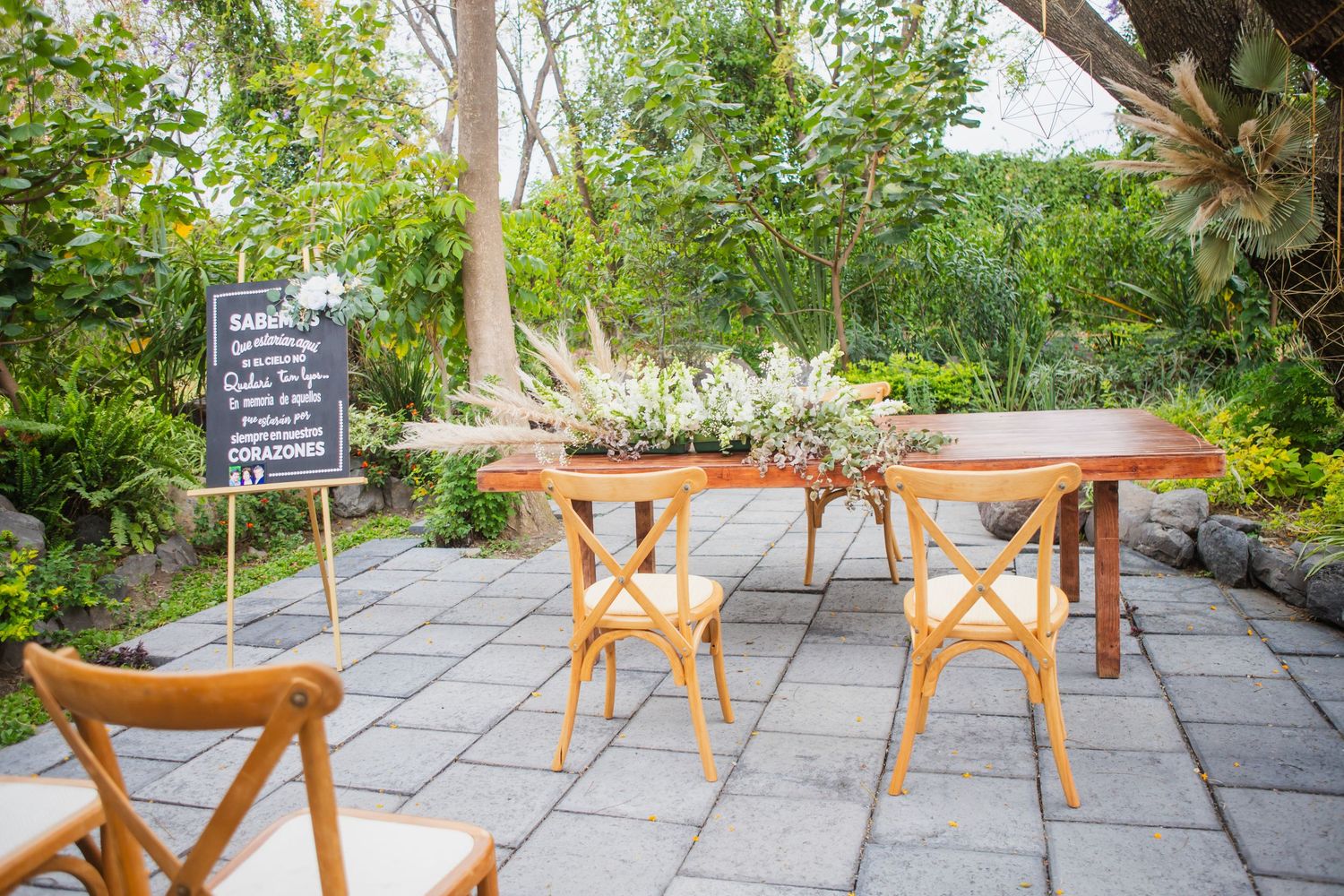 Rustic wooden table with floral centerpiece and wooden chairs on a stone patio beside a chalkboard sign in a lush garden wedding venue.