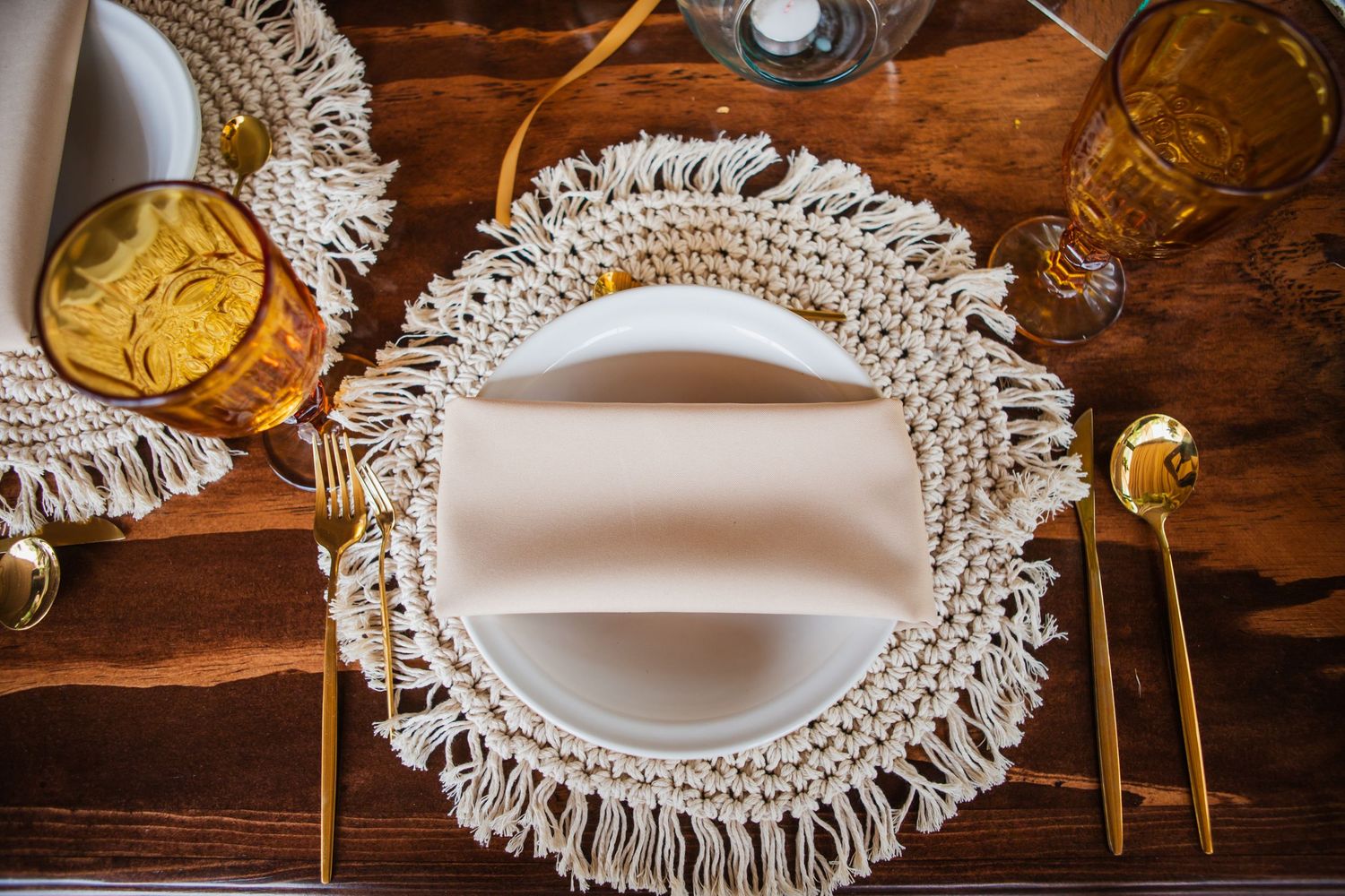 Boho table setting with a macrame fringe placemat, white plate, blush napkin, gold flatware and amber glassware on a rustic wooden table.