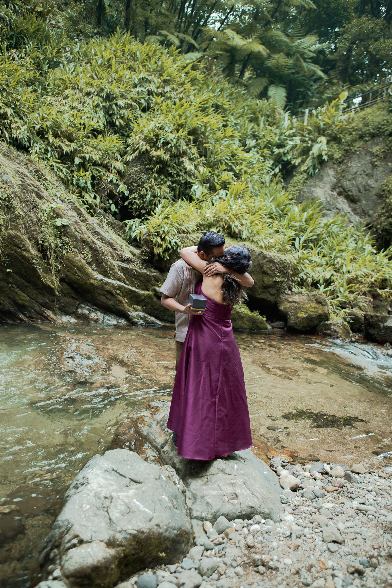 Couple embracing on a large rock in a clear forest stream, the woman in a flowing magenta dress, surrounded by lush green foliage.