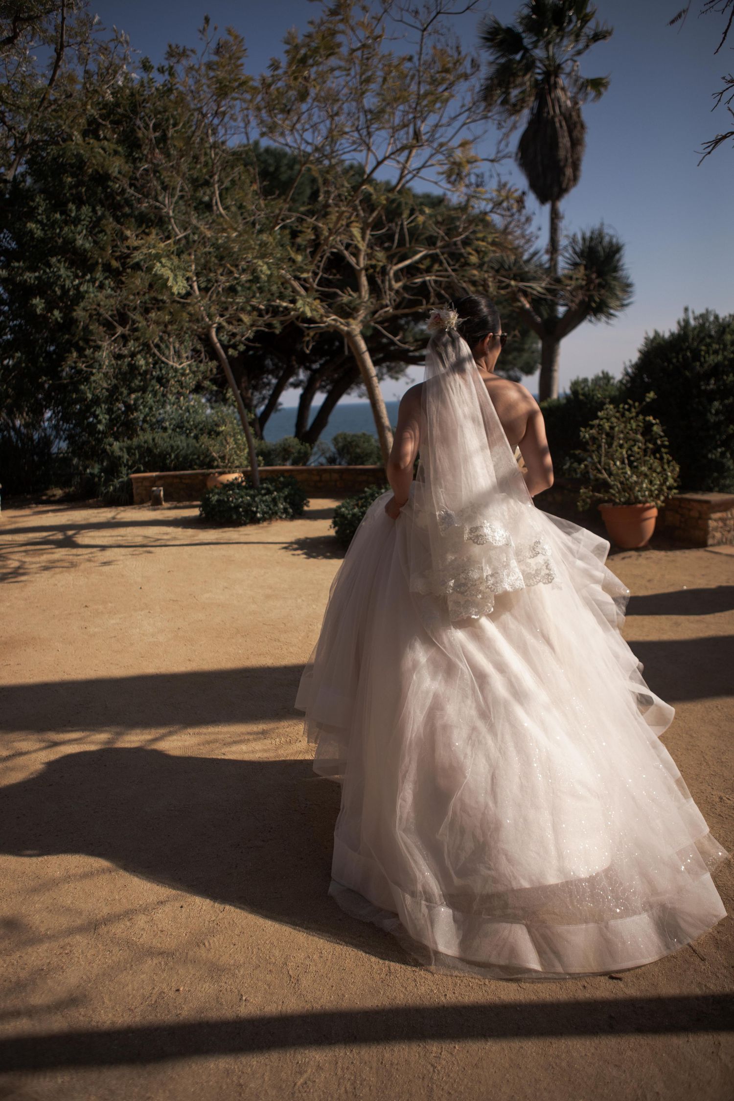 Bride in a voluminous tulle ball gown and long veil walks along a sunlit terrace shaded by trees.