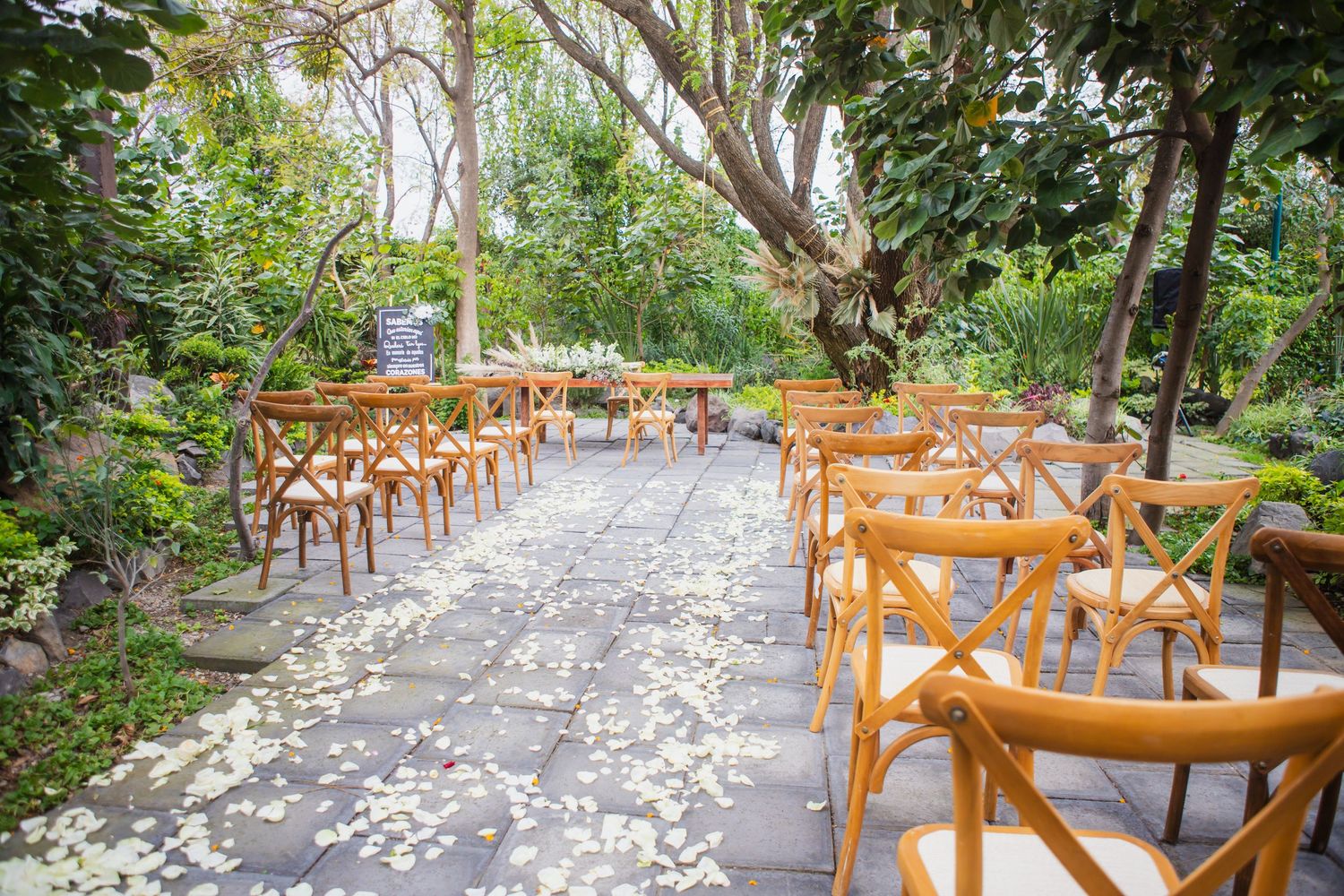 Sillas de madera junto a un pasillo de piedra cubierto de pétalos blancos, preparado para una ceremonia íntima en un jardín.