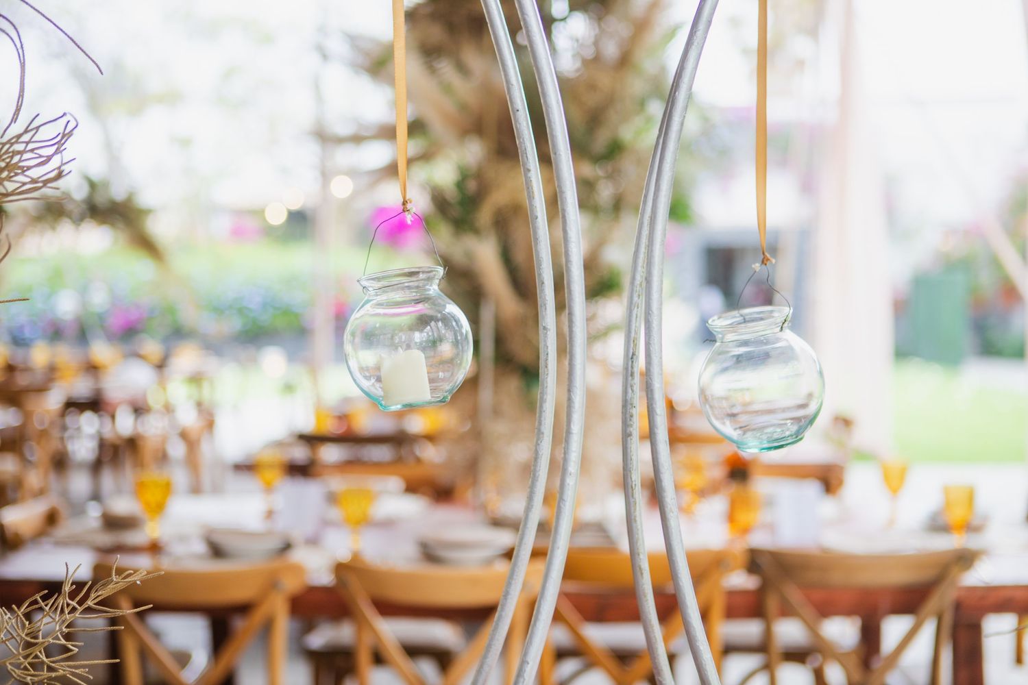 Glass hanging tea light holders on curved metal stands at an elegant outdoor wedding reception with wooden tables and chairs.
