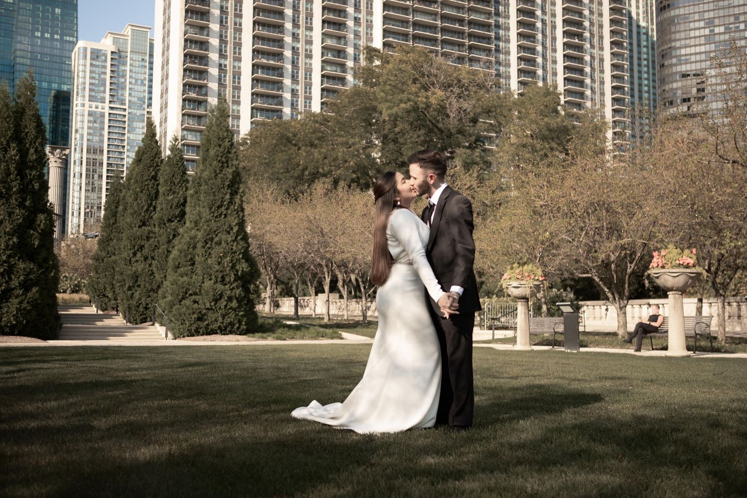 Newlywed couple kisses on a park lawn with tall city skyscrapers and trees in the background.