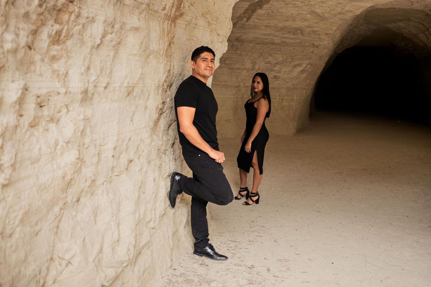 Couple in black outfits posing in a sandy limestone tunnel, the man leaning on the wall while the woman stands behind him.
