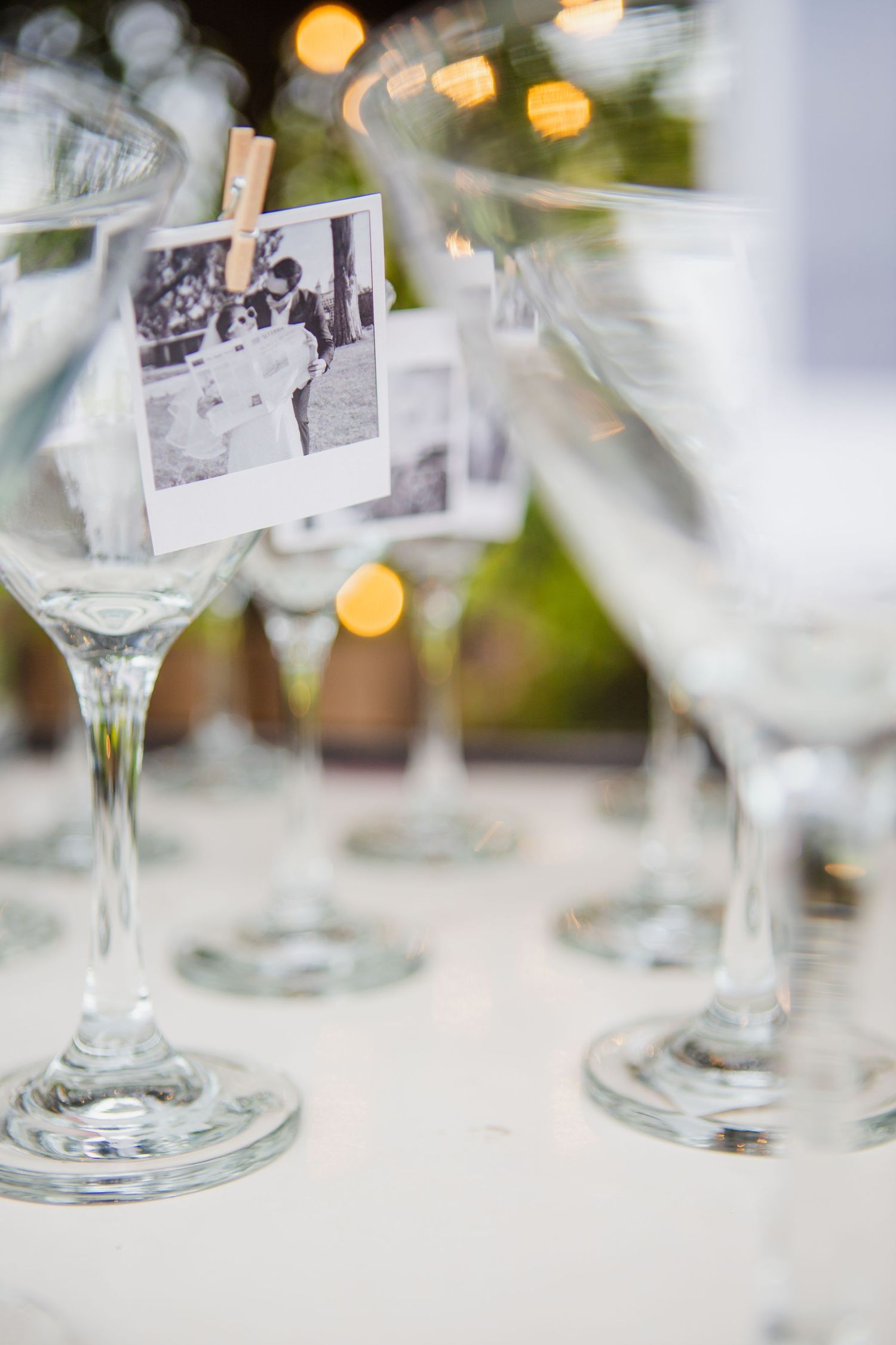 Black and white instant photo clipped with a mini wooden clothespin to a clear stemmed glass, with more glasses and warm string lights behin
