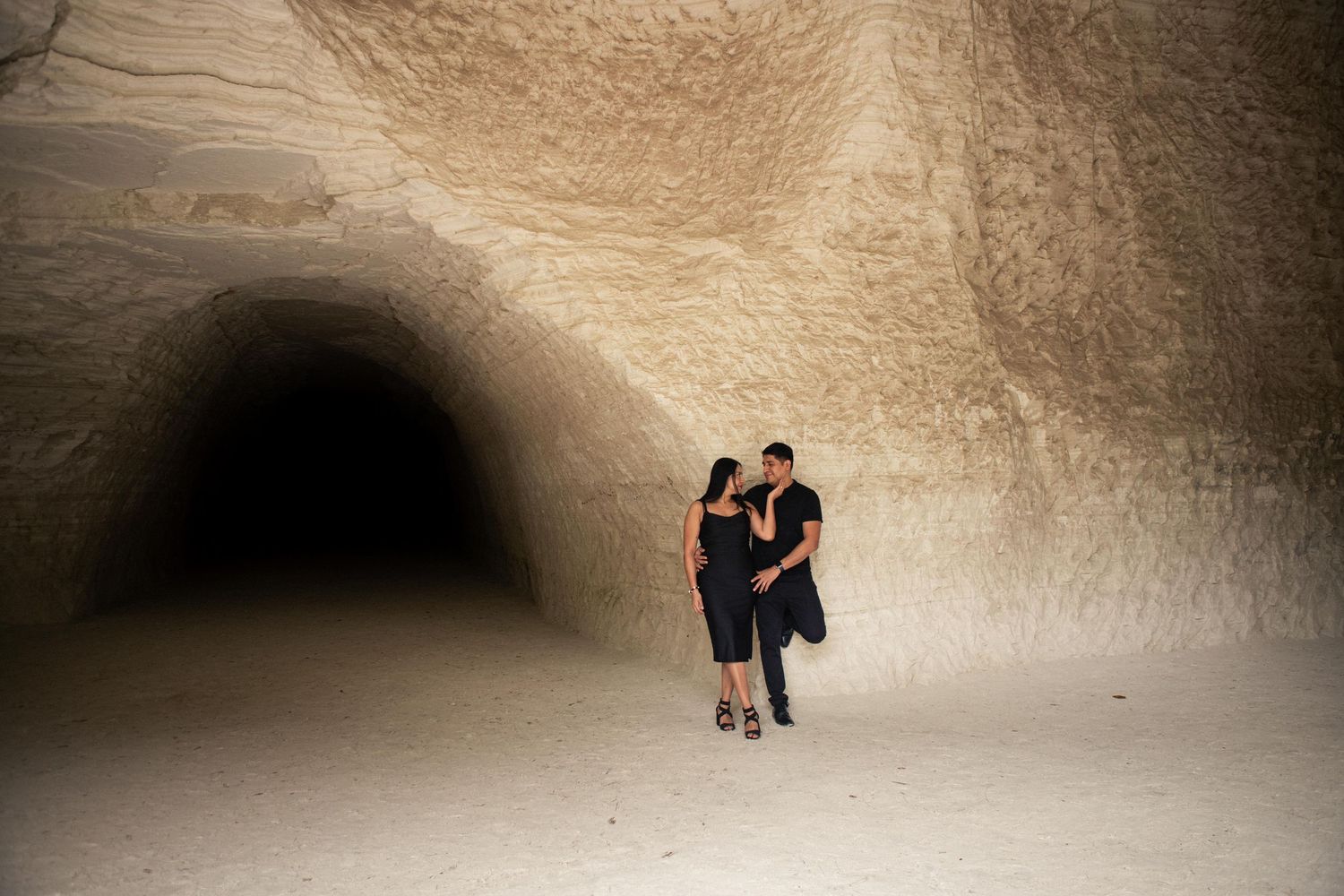 Couple in dark clothing stand close together in a large sandstone cave with smooth carved walls and a dark tunnel entrance behind them.