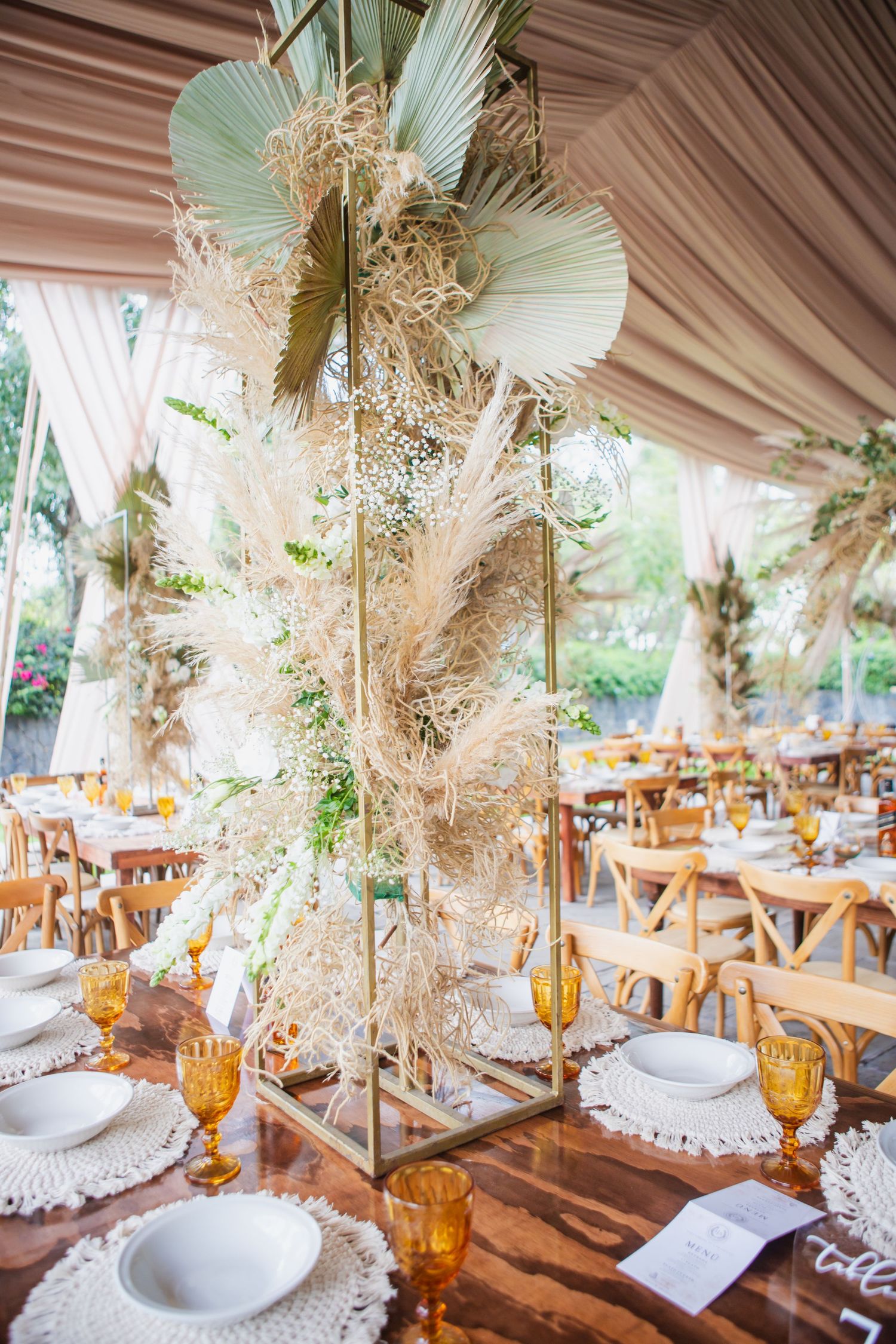 Tall gold frame centerpiece with pampas grass, dried palm leaves, and baby's breath on a wooden table at a boho tent wedding reception.