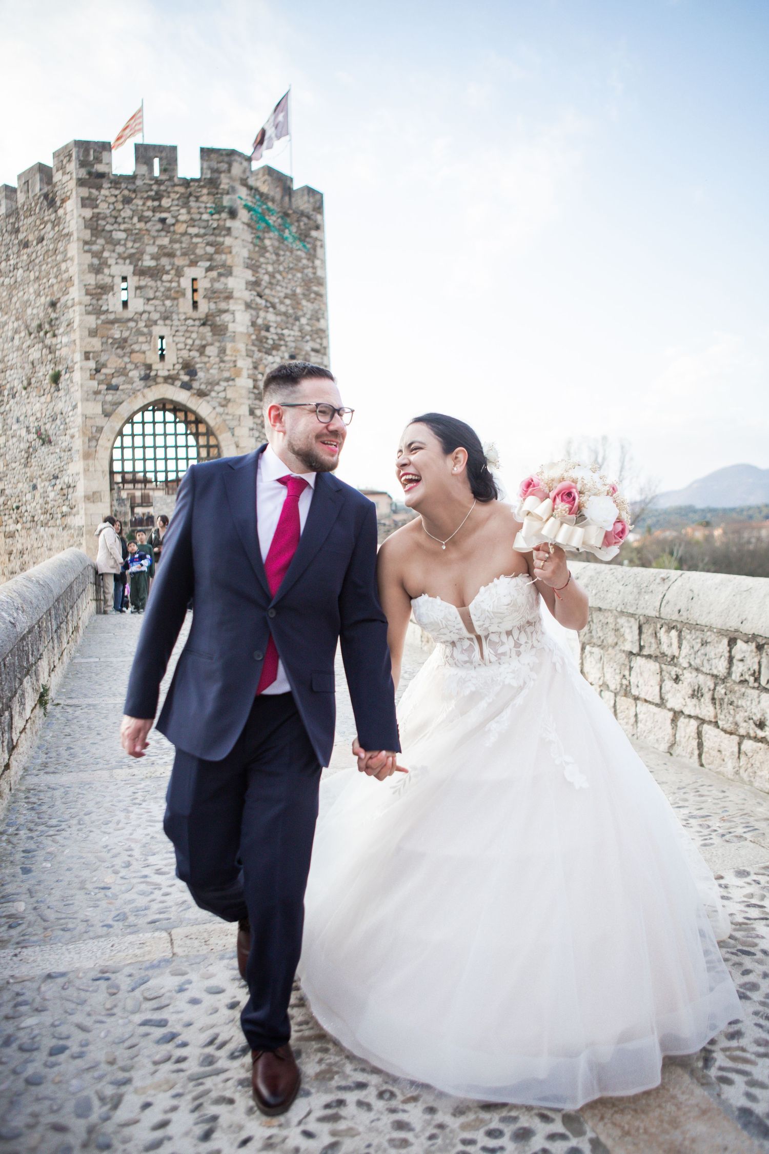 Laughing bride in a white gown with pink bouquet walks hand in hand with groom in navy suit across a stone bridge near a medieval tower.