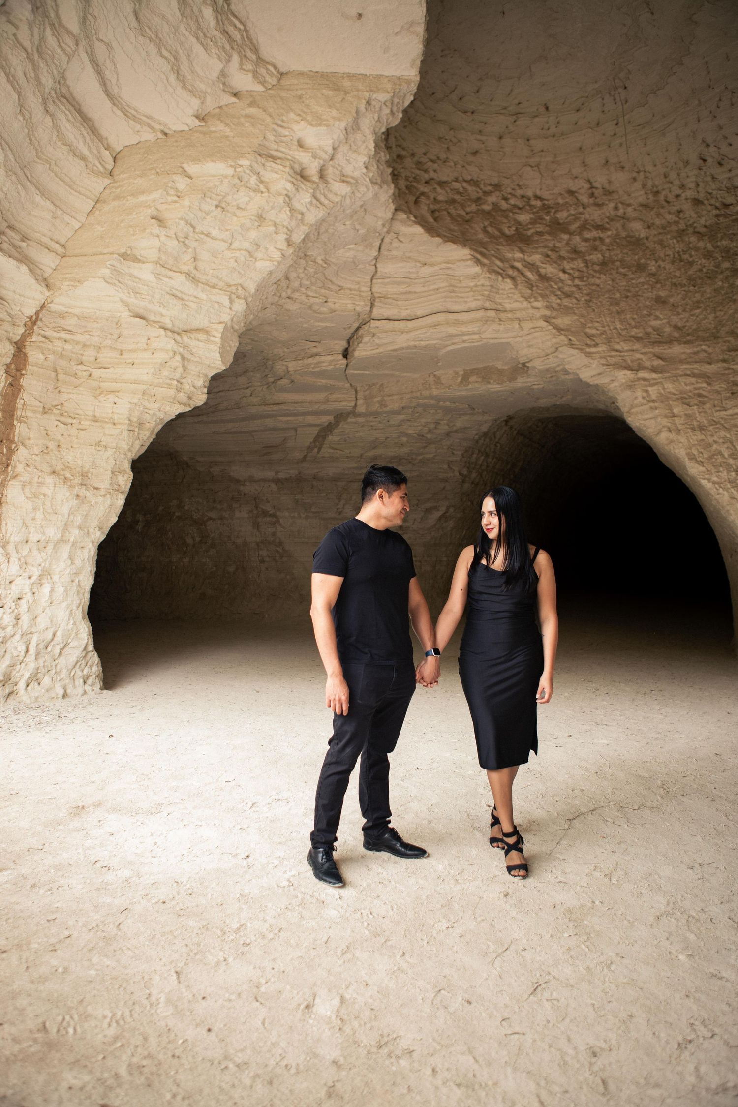 Couple in black outfits holding hands and smiling at each other in front of a large sandstone cave entrance for an engagement photo shoot.