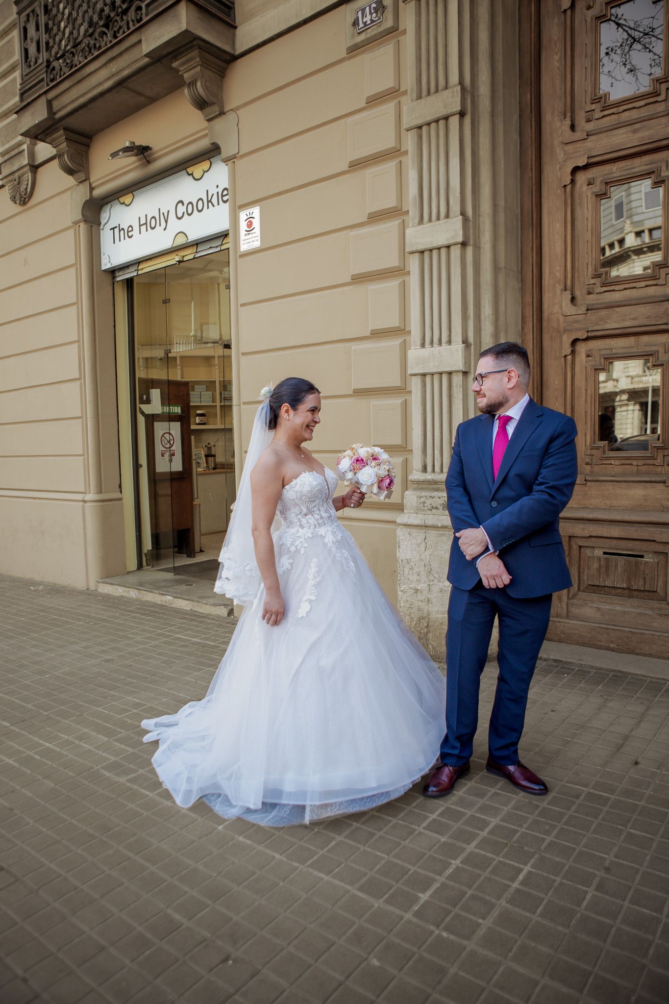 A bride in a white tulle gown holding a pink bouquet smiles beside a groom in a blue suit and pink tie outside The Holy Cookie shop.