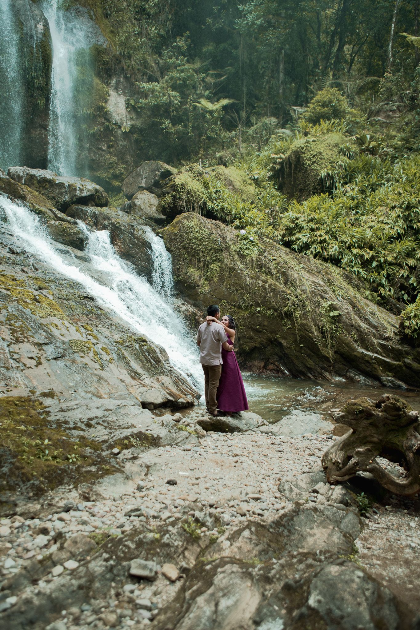 Couple in an embrace on rocky ground beside a cascading waterfall surrounded by lush green forest.