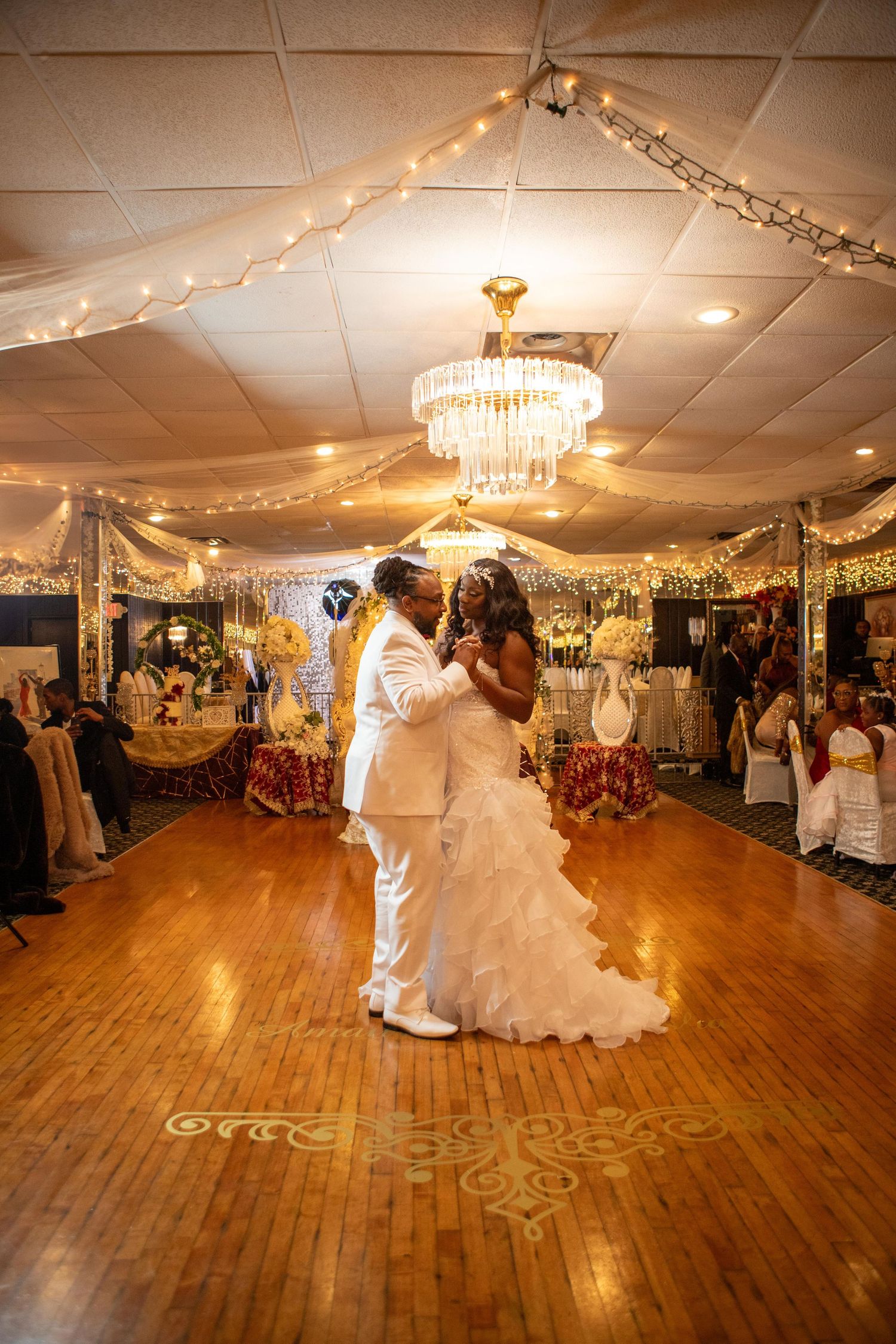 Bride in a white ruffled gown and groom in a white suit share a first dance on a wood floor under a chandelier at a wedding reception.