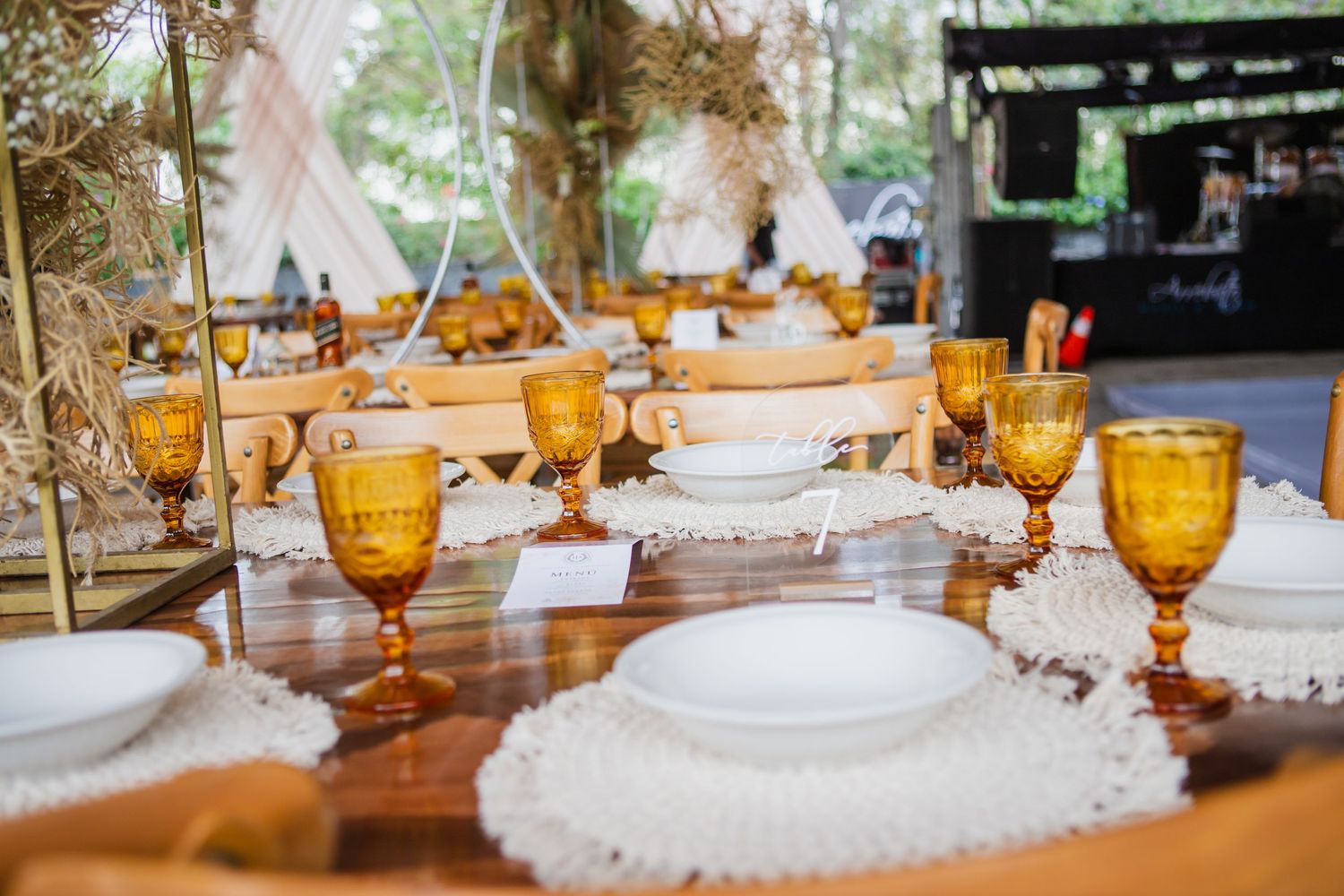 Rustic wooden table set with amber glass goblets, white plates and crochet lace placemats for an elegant outdoor boho wedding reception.