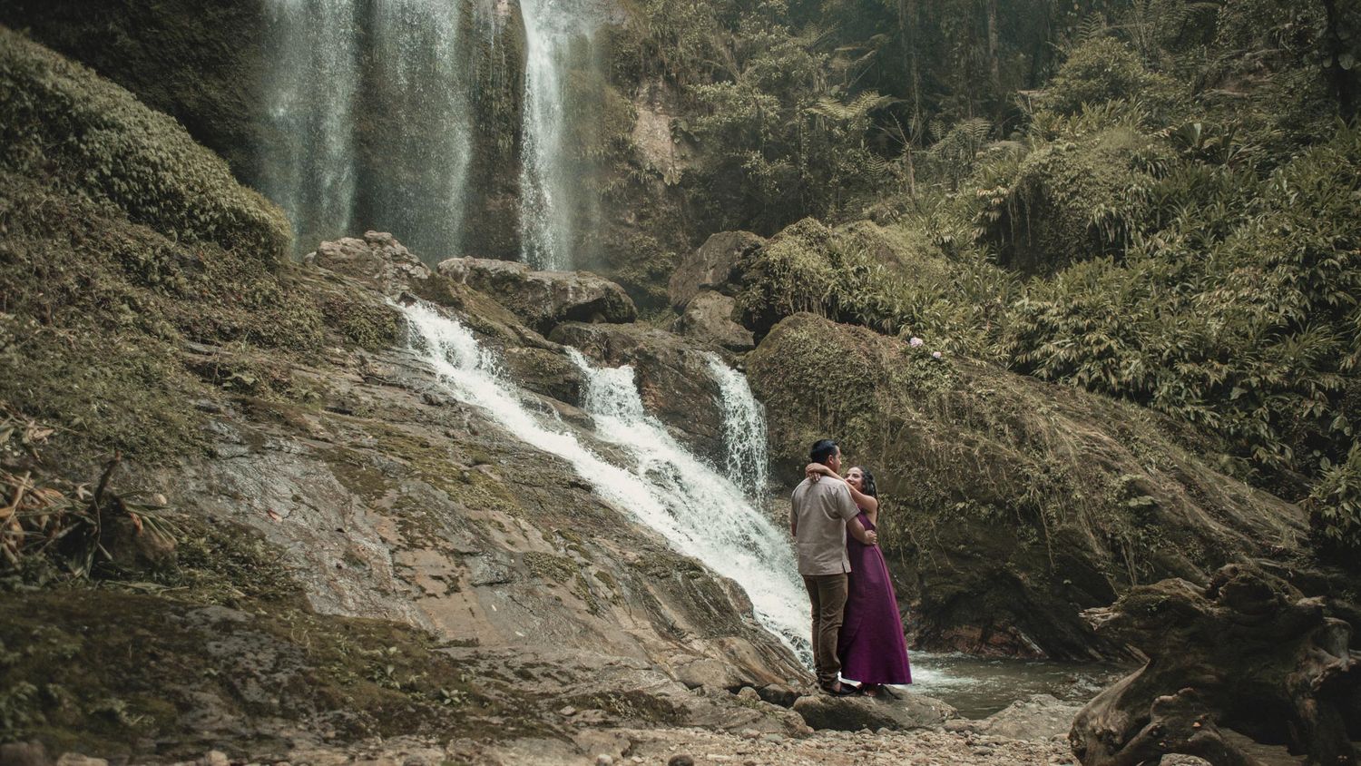 Couple in a purple dress embracing beside a cascading waterfall surrounded by mossy rocks and lush green forest.