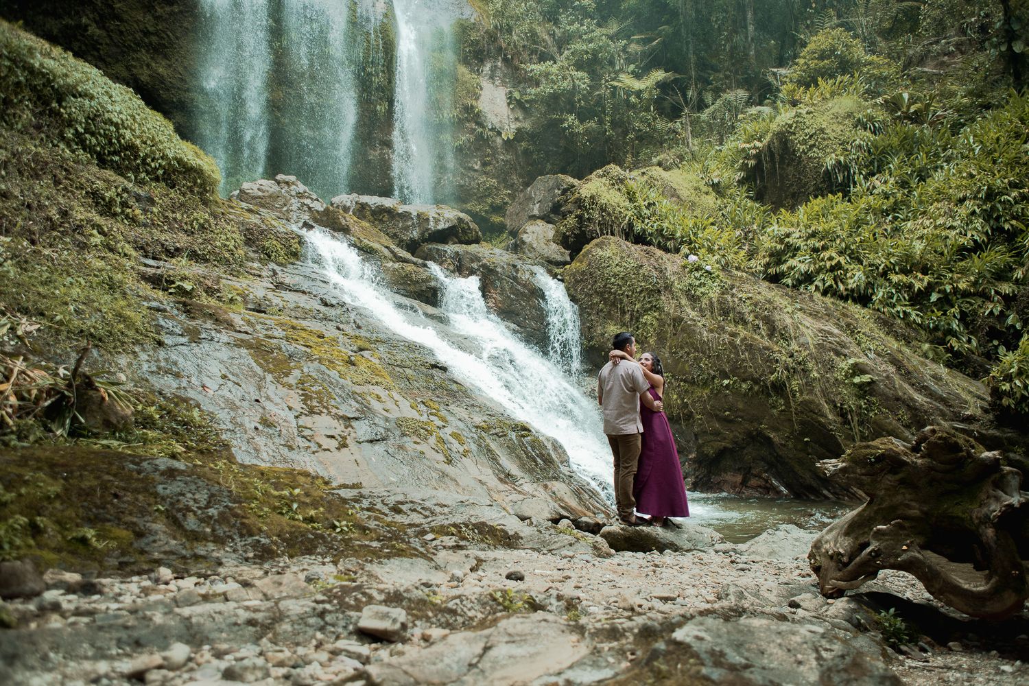 Couple embracing on mossy rocks beside a cascading waterfall surrounded by lush green forest, with the woman wearing a flowing purple dress.