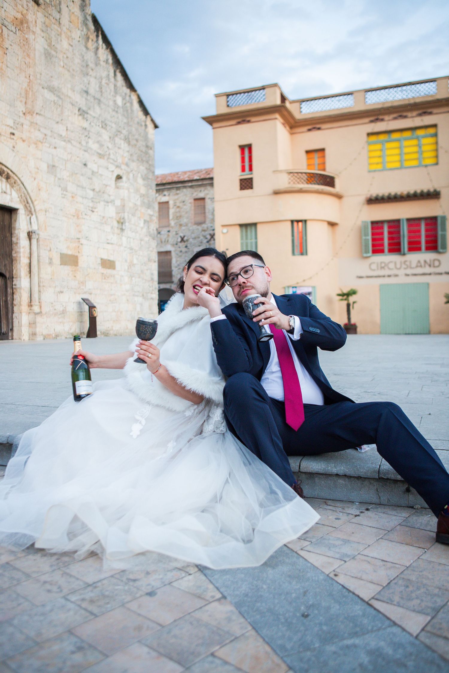 Bride in a white gown and groom in a navy suit sit on stone steps in a historic plaza, holding wine glasses and a champagne bottle.