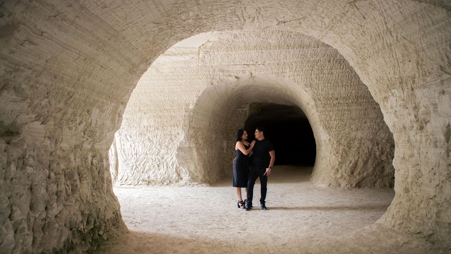 Couple embracing in a sandy limestone cave tunnel with smooth carved arches leading toward a dark passage.