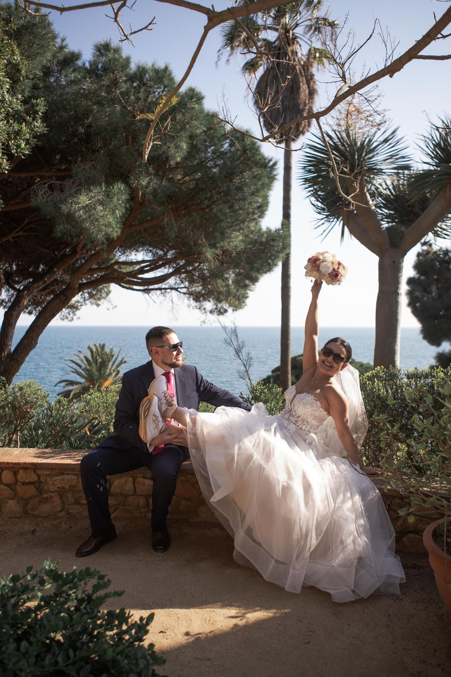 Groom in a dark suit sits on a stone wall as the bride raises her bouquet, with the sparkling sea and palm trees behind them.