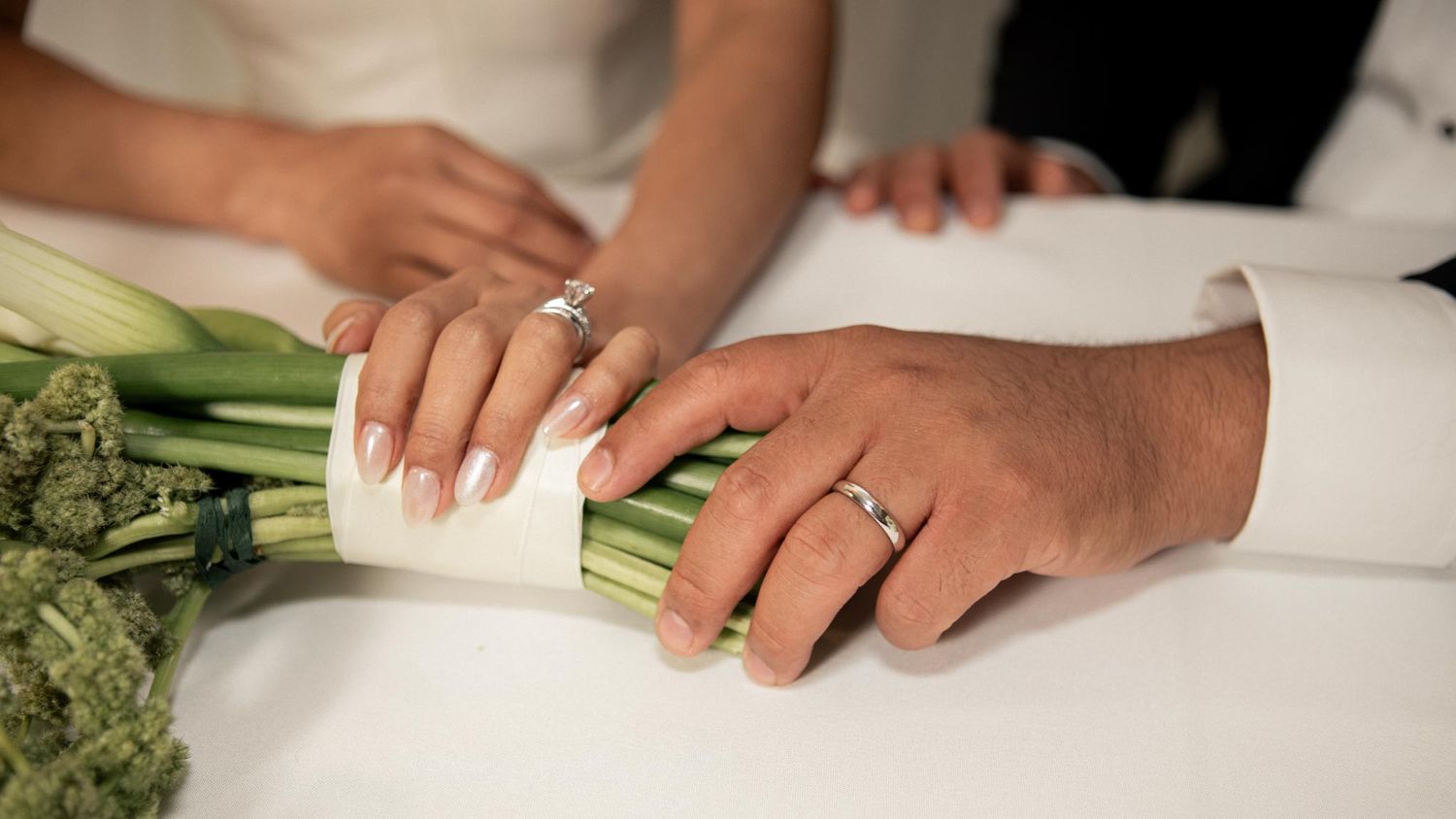 Detalle de las manos de los novios con sus anillos de boda sobre un ramo de flores blancas.