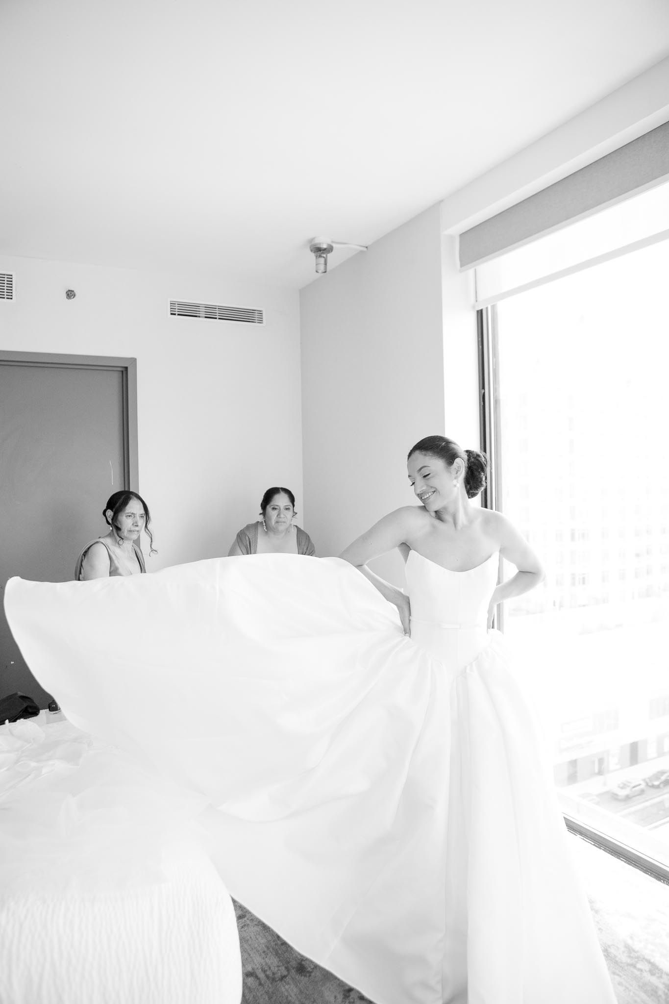Smiling bride spreads out the skirt of her white wedding gown while two bridesmaids watch in a black and white photo.