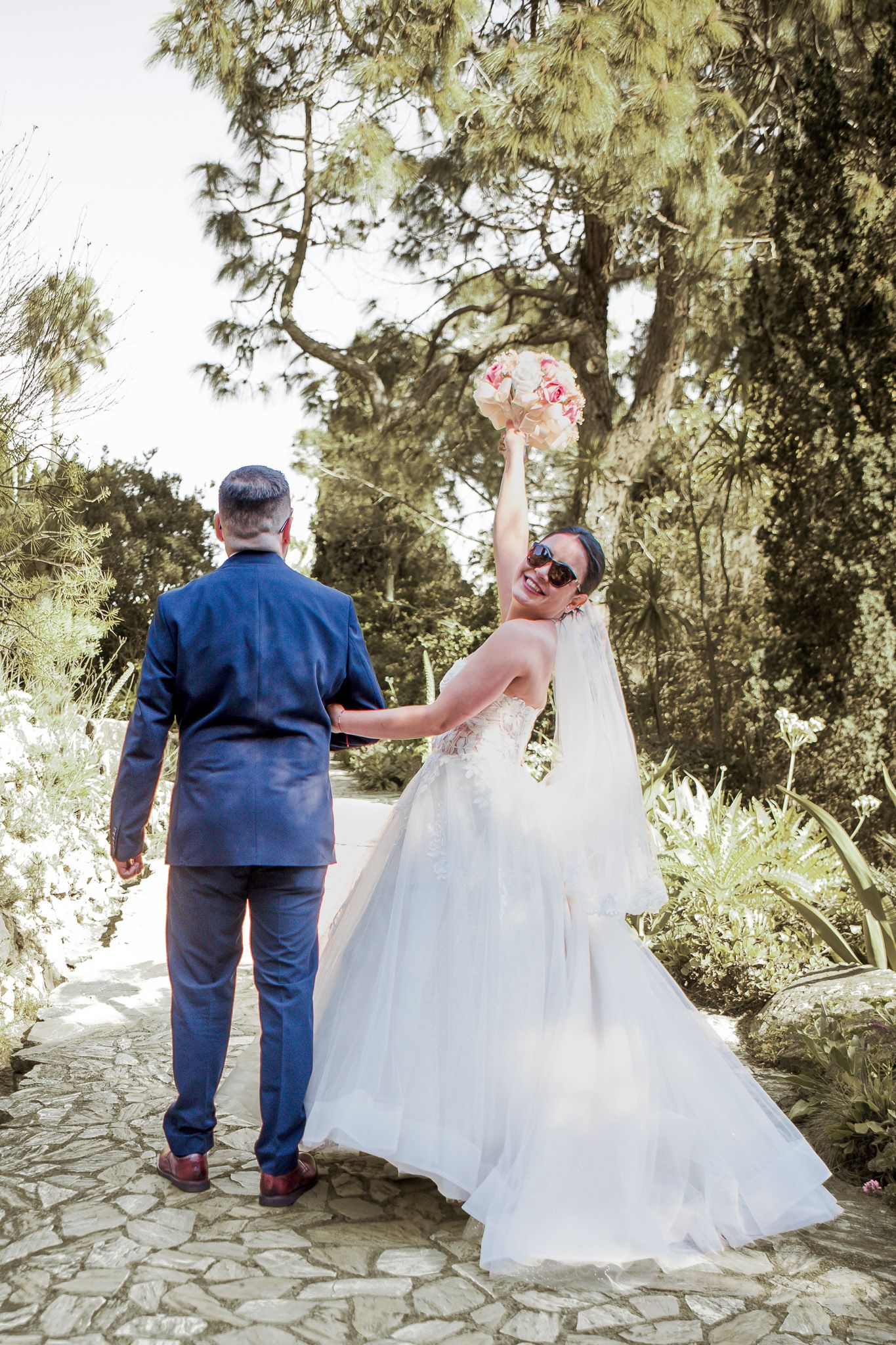 Bride in a flowing white gown and sunglasses raises her bouquet joyfully beside a groom in a blue suit on a stone garden path.