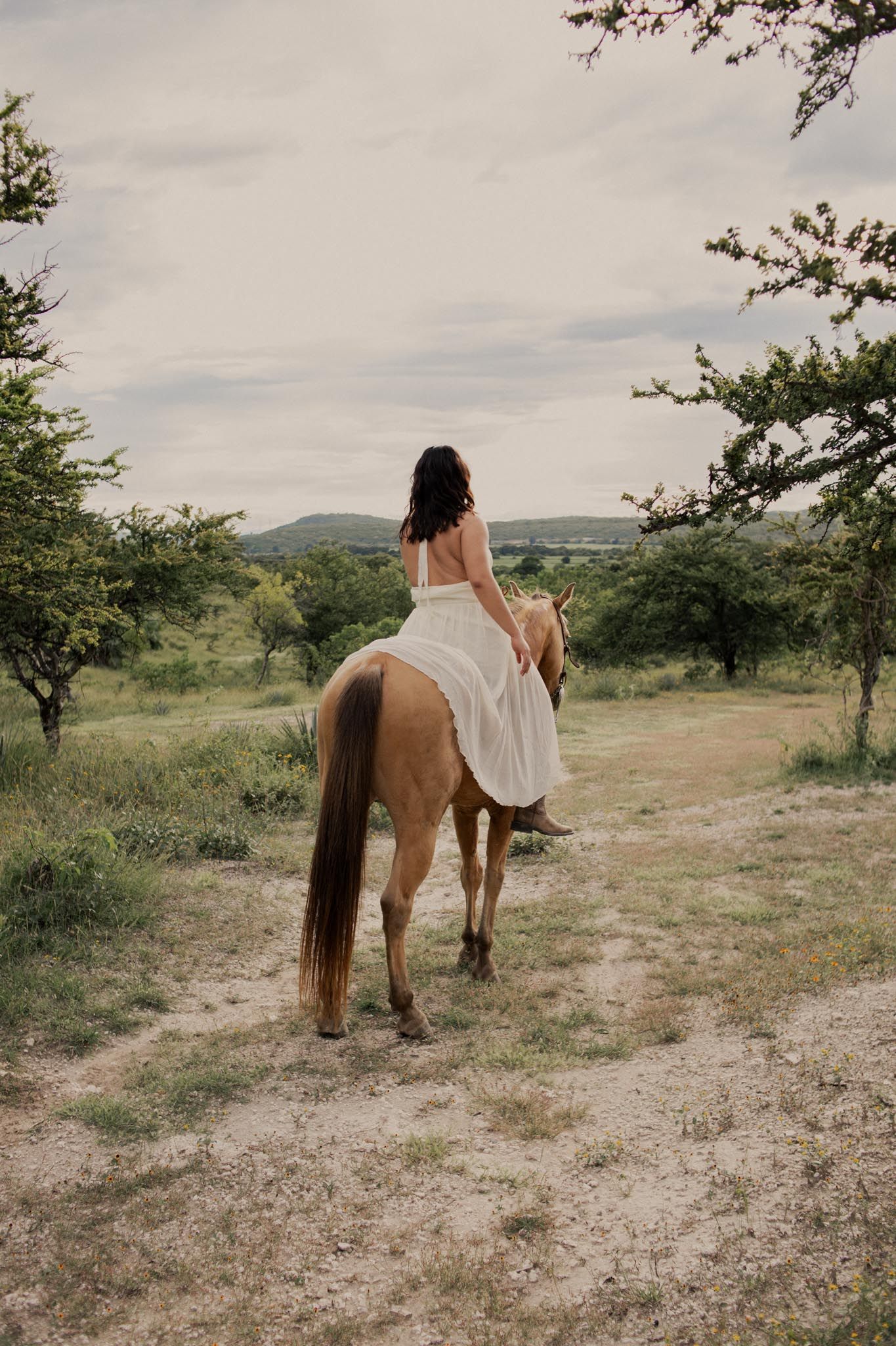 Woman in a flowing white dress rides a chestnut horse down a dirt path through green countryside under a cloudy sky.