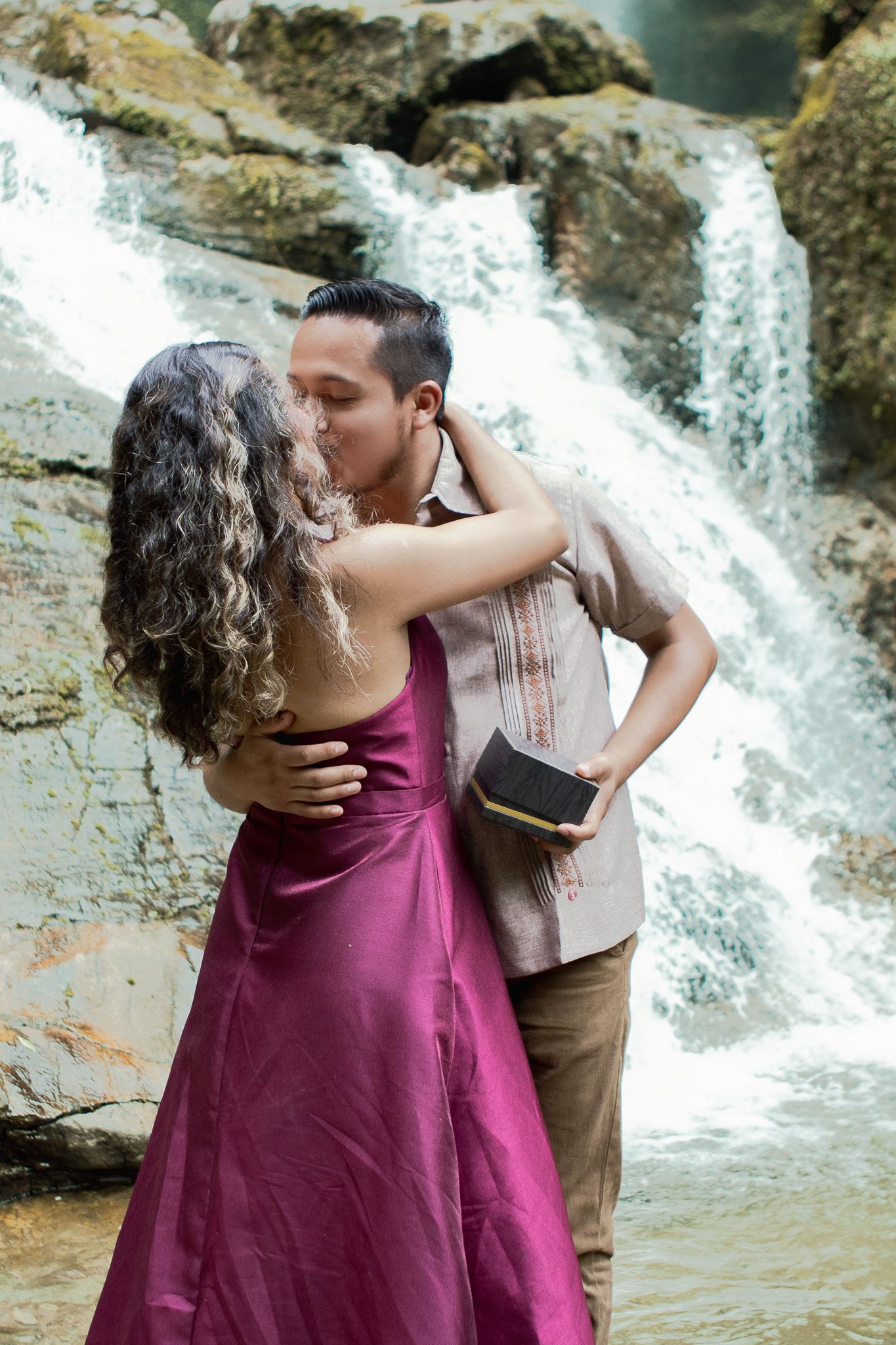 Couple kissing and embracing in front of a waterfall, the woman in a magenta satin gown and the man holding a small book.