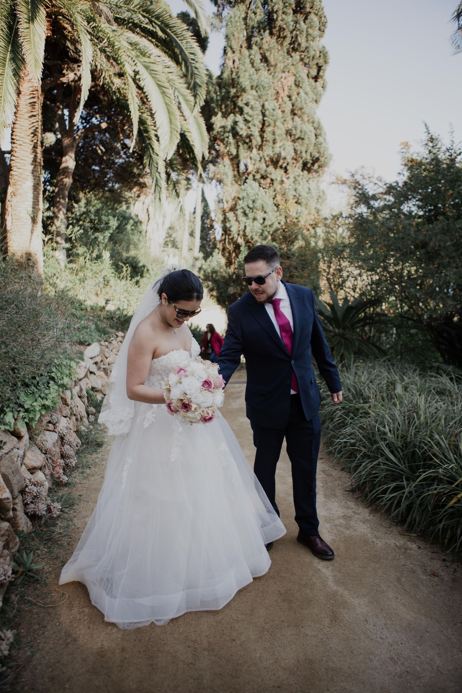 Novios caminando de la mano por un sendero ajardinado en la Costa Brava, ella con ramo blanco y él con corbata rosa.