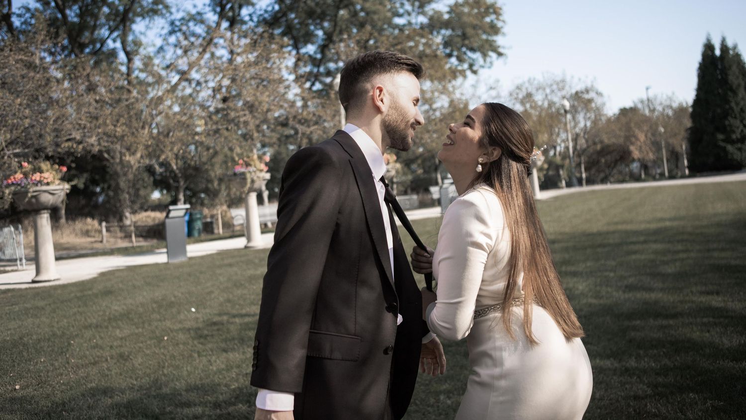 Pareja sonriendo frente a frente en un parque, él con traje oscuro y ella con vestido blanco de manga larga.