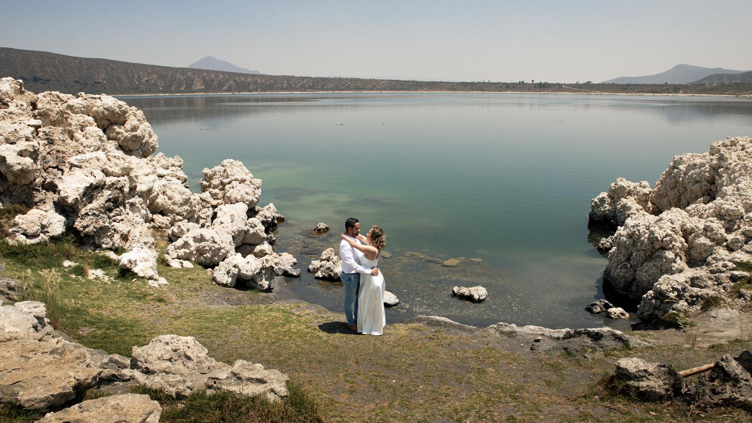 Couple in wedding attire embracing on a grassy shore beside a calm lake, with white rock formations and distant mountains under a clear sky.