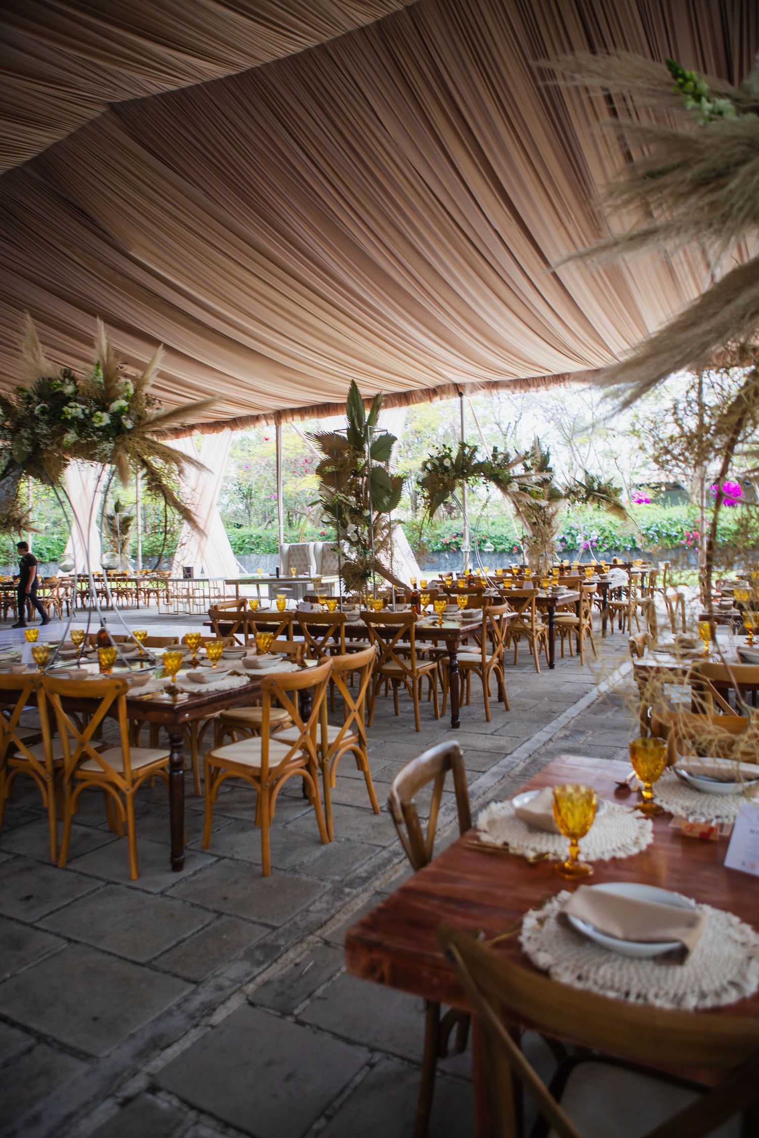 Elegant outdoor wedding reception under draped tent with wooden tables, cross-back chairs, woven placemats, amber glassware and tropical flo