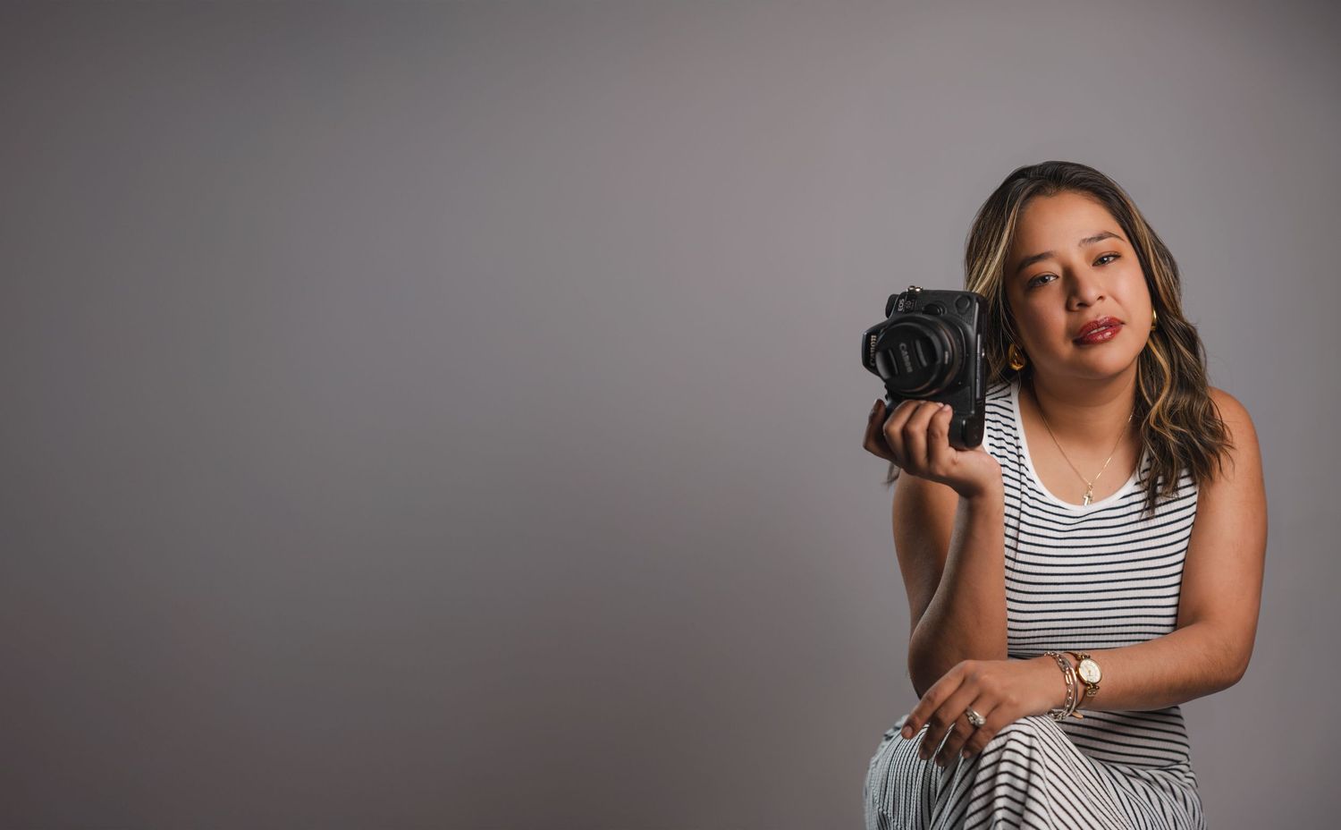 Woman with long wavy hair in a striped sleeveless top holds a black camera while posing against a gray studio backdrop.