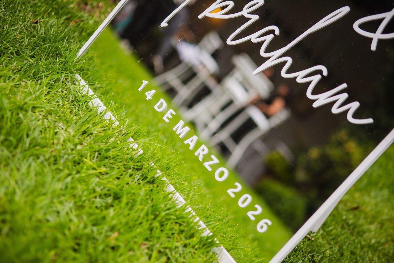 Mirrored wedding sign with white script lettering and the date 14 de marzo 2026 resting on green grass, with white chairs blurred behind.