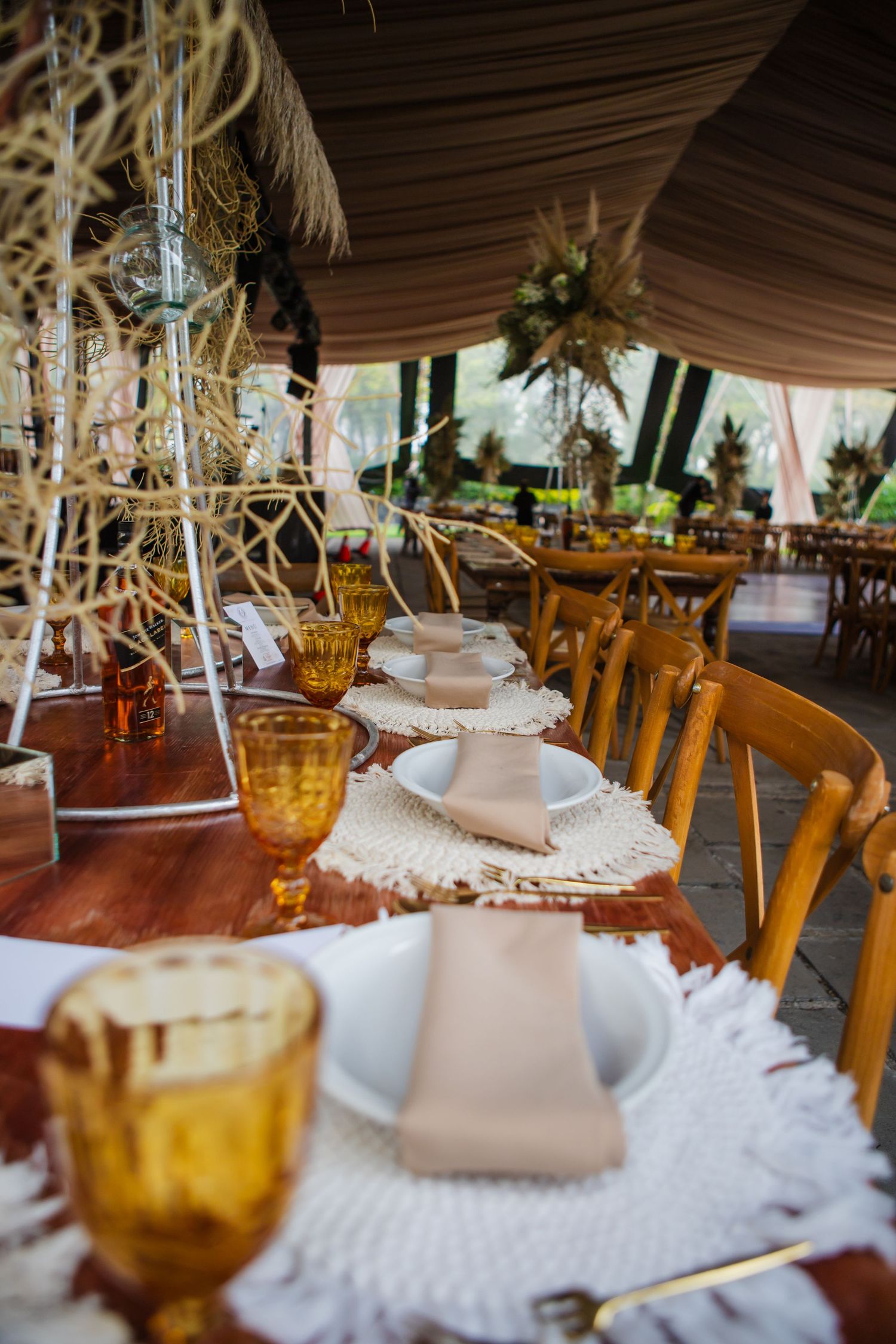 Boho wedding table setting with amber goblets, woven fringe placemats, blush napkins on white plates, and wooden chairs under a draped tent.