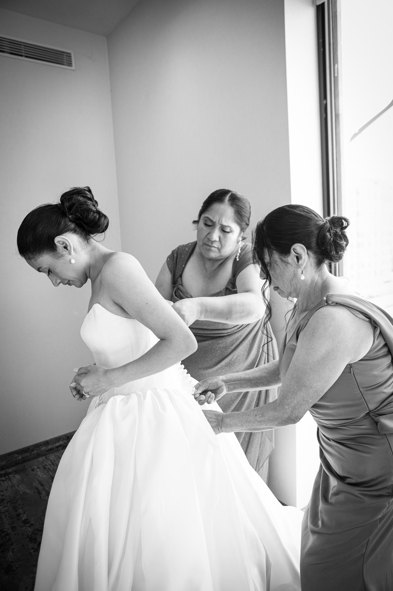 Two women help the bride button and adjust her strapless wedding gown in a black and white getting-ready photo.