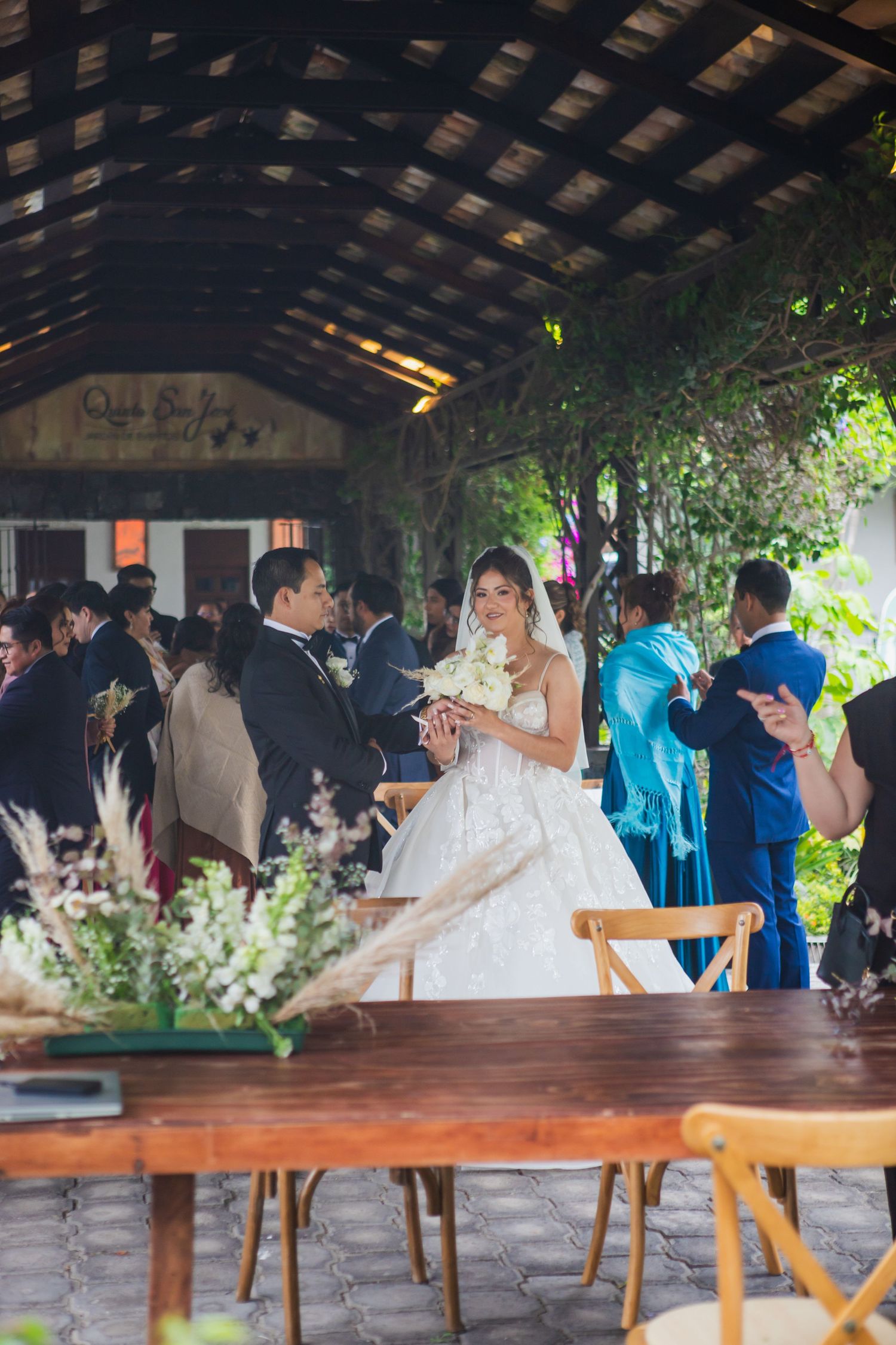 Bride in a white gown holding a bouquet walks past a rustic wooden table with floral centerpieces as wedding guests mingle under a pergola.