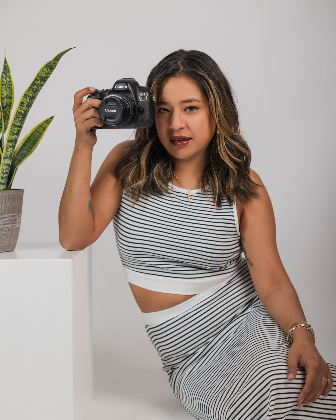 Woman in a striped sleeveless top and skirt sits beside a white pedestal holding a camera next to a potted snake plant.