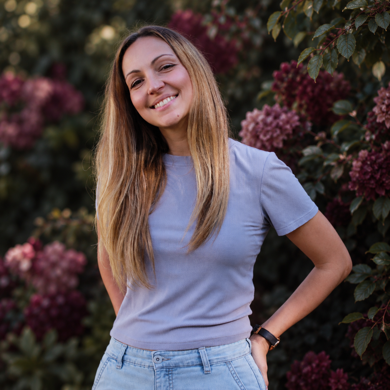 Smiling young woman with long blonde hair in a grey t-shirt posing near pink flowering bushes.