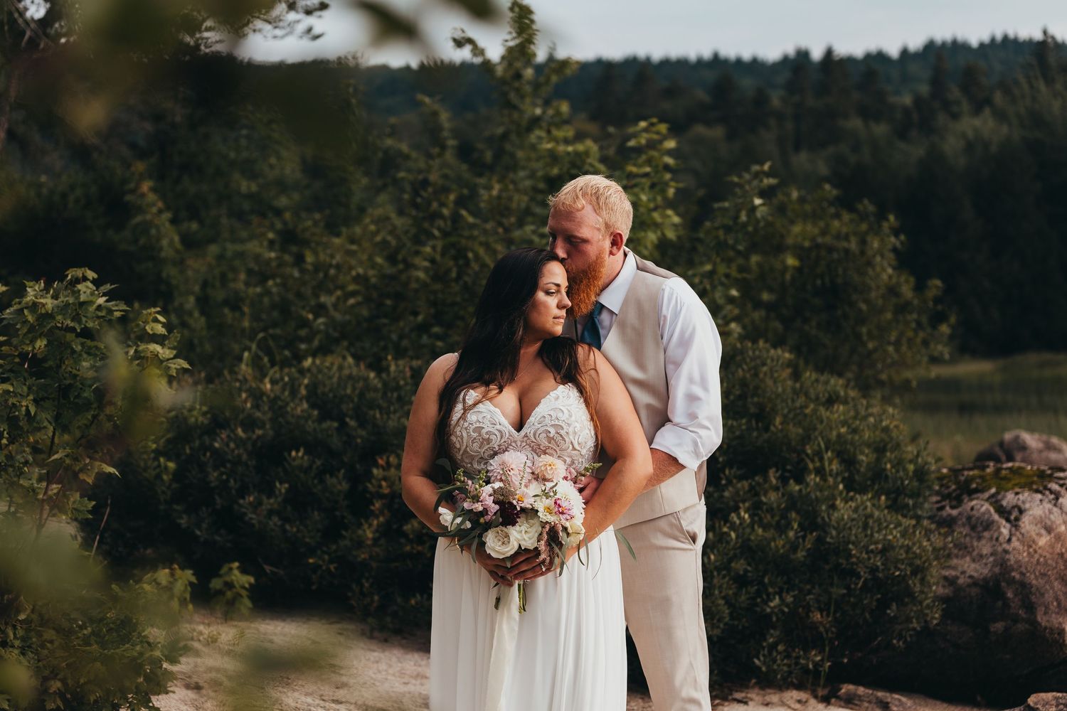 Bride and groom embrace outdoors, she holds a floral bouquet, surrounded by lush green trees at a forest wedding.