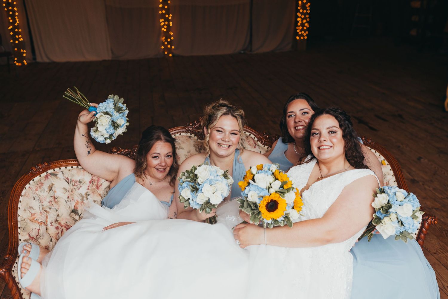Four bridesmaids in white and light blue dresses holding sunflower and blue flower bouquets seated on vintage sofa.