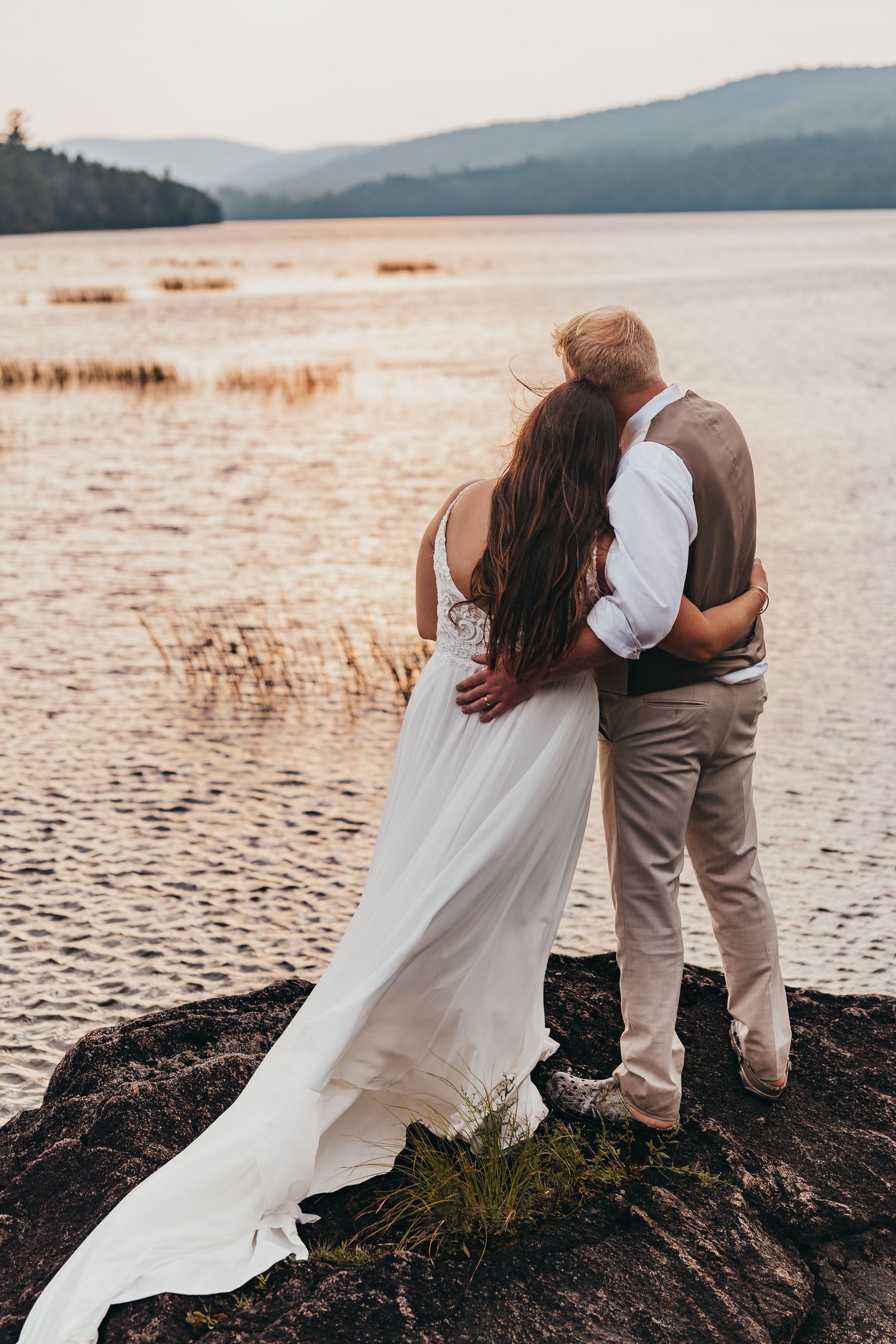 Couple embracing at sunset by a serene lake with mountains in the background.